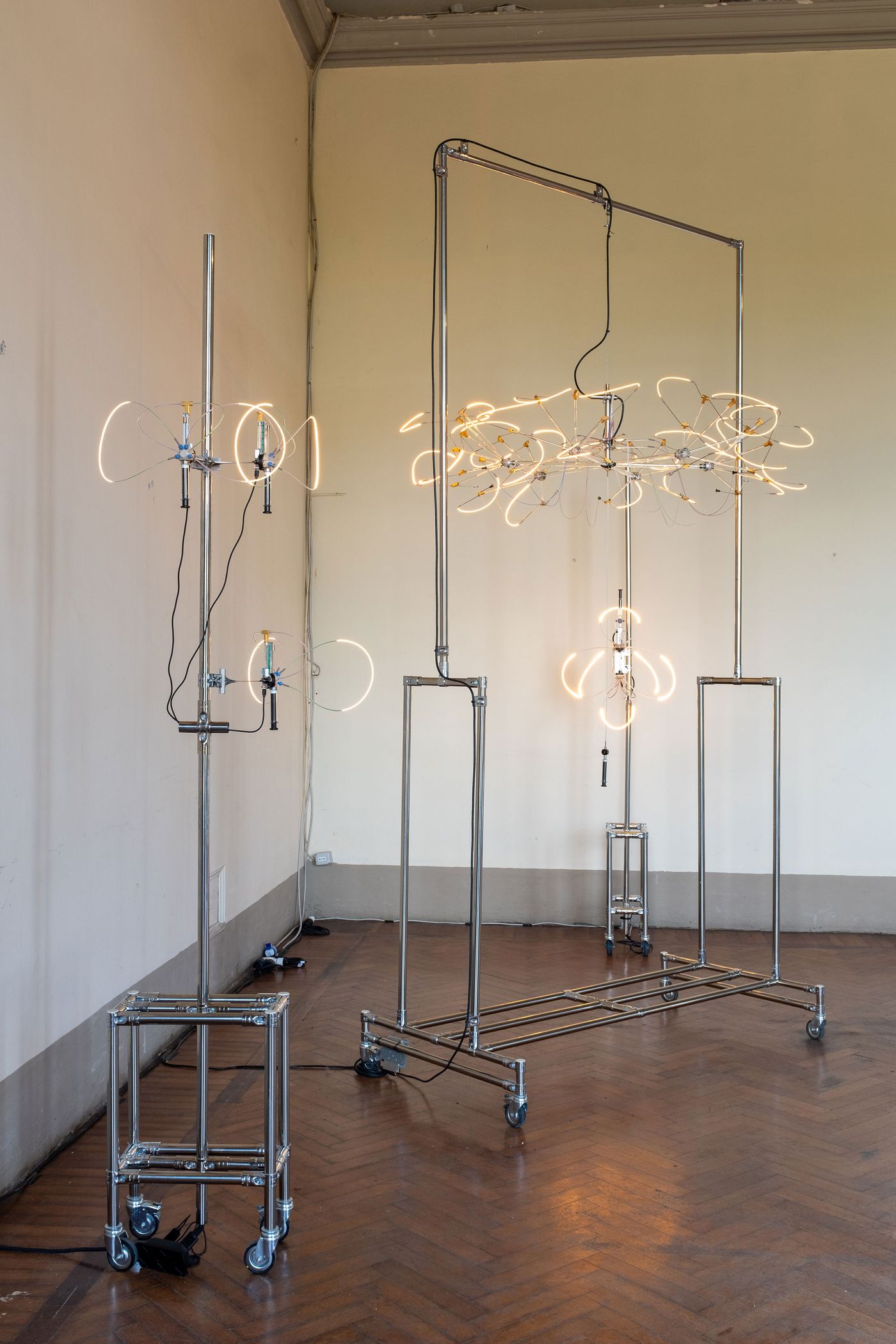 A freestanding sculptural light installation made of stainless steel tubing, glowing fiber-optic loops, and delicate wires on caster wheels, arranged within a minimalist room with herringbone wood floors.