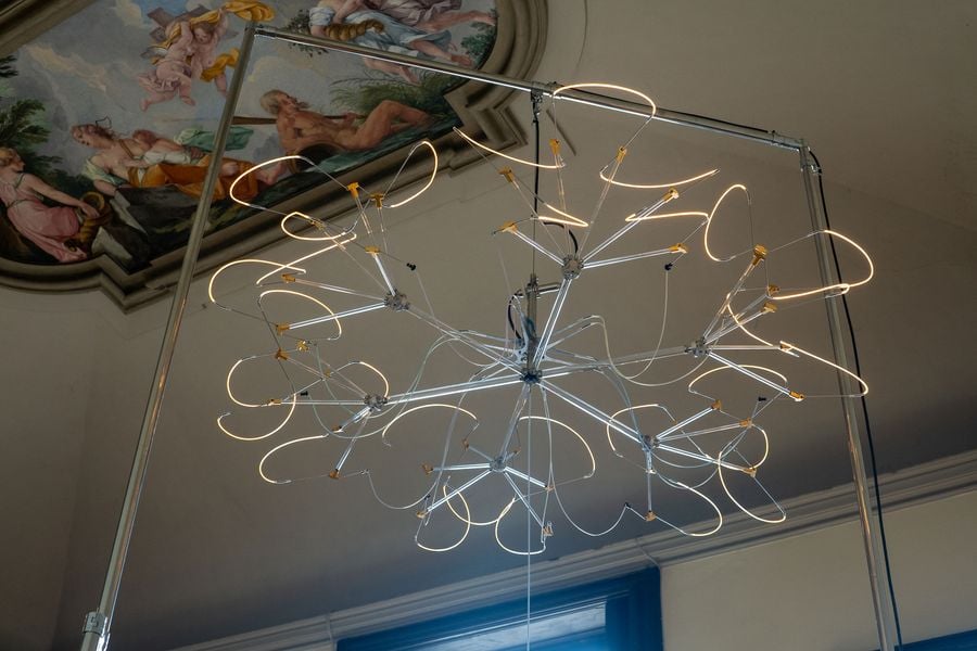A large chandelier-like installation made of glowing light loops arranged in a starburst pattern, suspended from a tall metal frame, with a frescoed ceiling in the background adding a contrast of classic and contemporary.