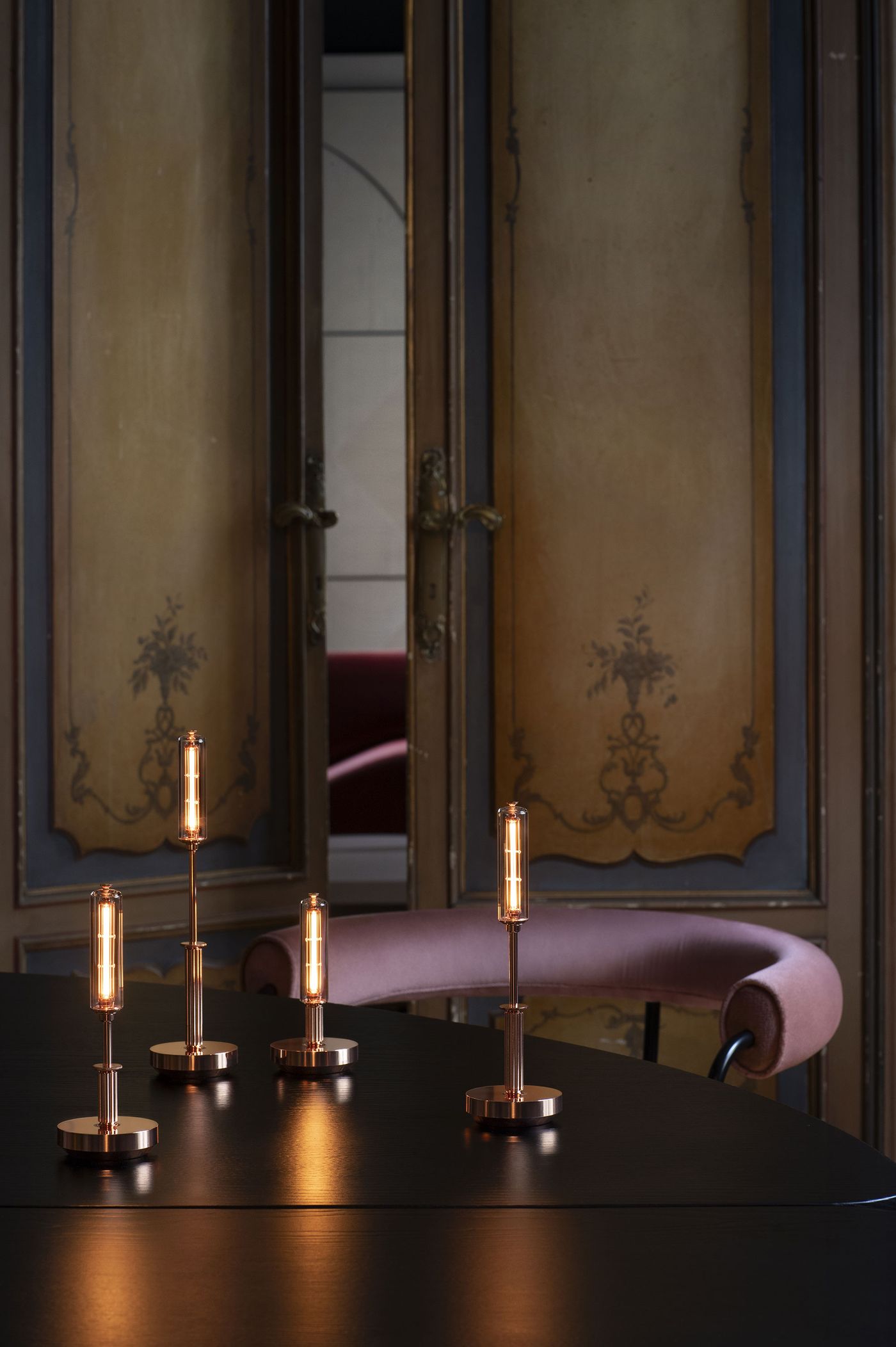 Four slim, cylindrical table lamps with glowing vertical filaments and metallic bases are arranged on a dark tabletop in front of ornate, vintage wooden doors. A mauve upholstered chair curves behind the table.