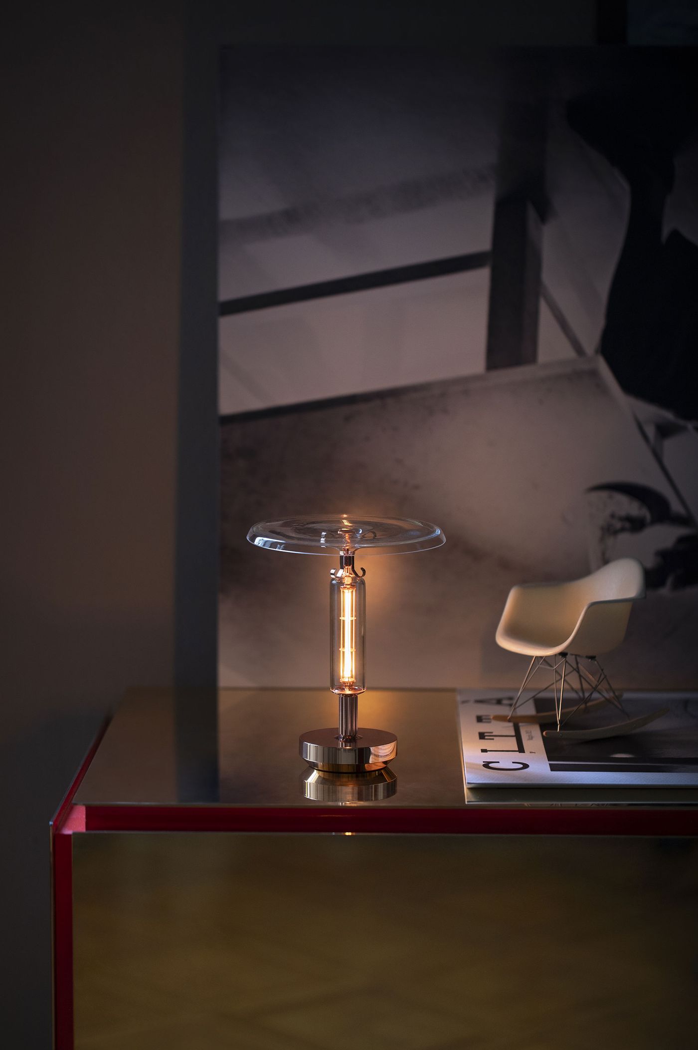 A single modern table lamp with a cylindrical glass body and warm filament stands on a reflective glass-topped table. The lamp features a wide, flat glass disc as a shade. In the background, a miniature white Eames-style chair sits on a magazine.