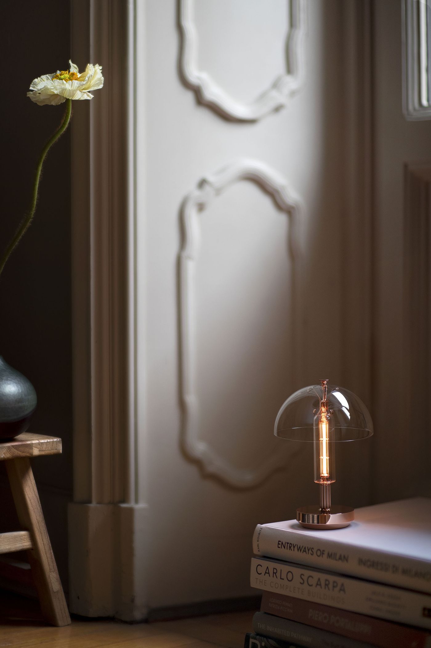 A small table lamp with a cylindrical glass body and glowing filament, topped by a clear dome-shaped shade, rests on a stack of design books near a window with decorative molding. A single pale flower in a vase is partially visible to the left.