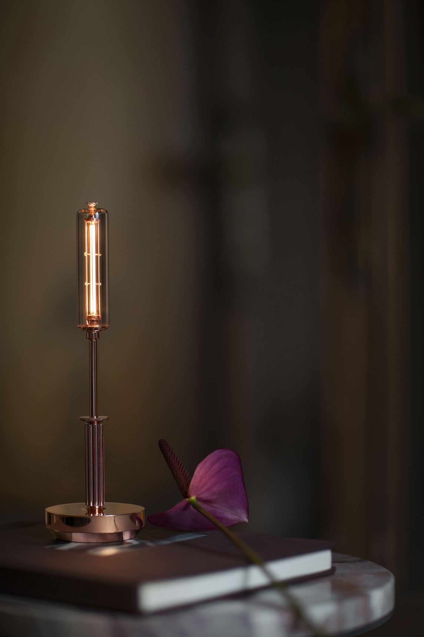 A close-up of a sleek, cylindrical table lamp with a glowing filament and metallic base sits on a round marble side table next to a pink anthurium flower and a closed notebook. The background is softly blurred.