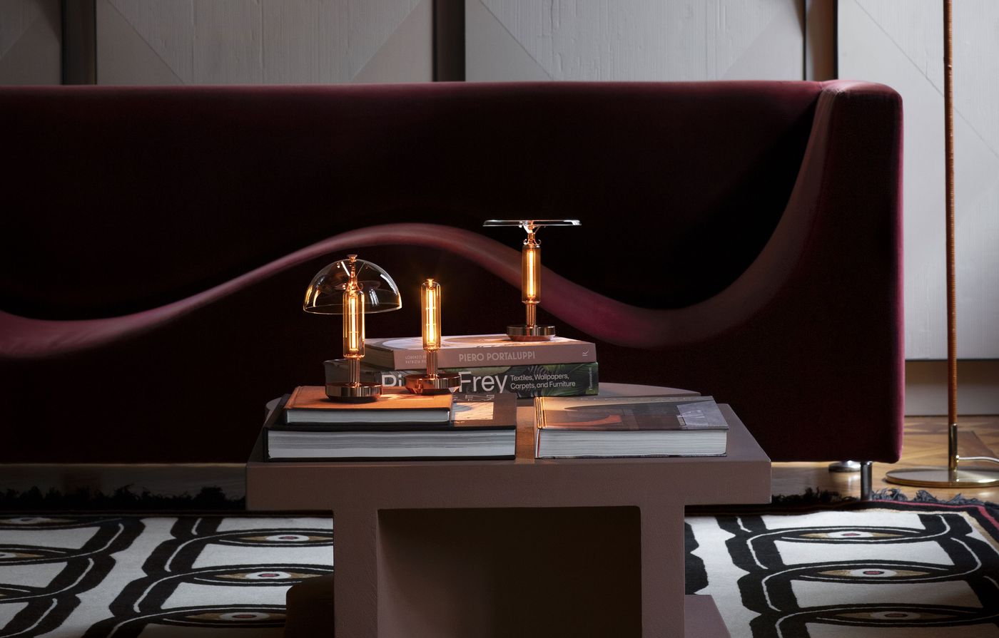 Three modern table lamps—one with a dome-shaped shade, one with a flat disc shade, and one with a simple cylindrical design—are arranged on a low coffee table stacked with art and design books. Behind them is a sculptural burgundy velvet sofa and a patterned rug. Three modern table lamps—one with a dome-shaped shade, one with a flat disc shade, and one with a simple cylindrical design—are arranged on a low coffee table stacked with art and design books. Behind them is a sculptural burgundy velvet sofa and a patterned rug.