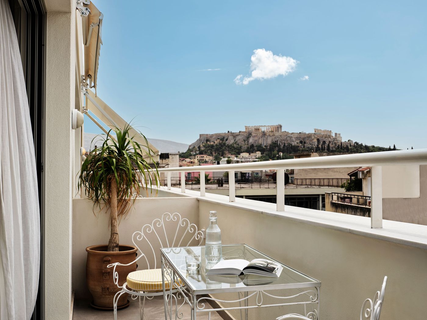A private balcony at Apollo Palm Hotel in Athens offers unobstructed views of the Acropolis and Parthenon against a clear blue sky. White wrought-iron chairs with yellow-striped cushions surround a glass-topped table, accompanied by a potted tree and an open book, creating an intimate Mediterranean setting framed by the city's rooftops.