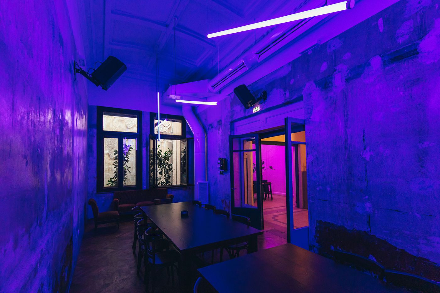A moody, industrial-style room at Arcade in Thessaloniki, bathed in electric purple and blue lighting. Distressed plaster walls, minimalist tables, dark wooden chairs and suspended LED light bars create an atmospheric, nightclub-like setting, while tall windows and glazed doors reveal adjacent spaces.