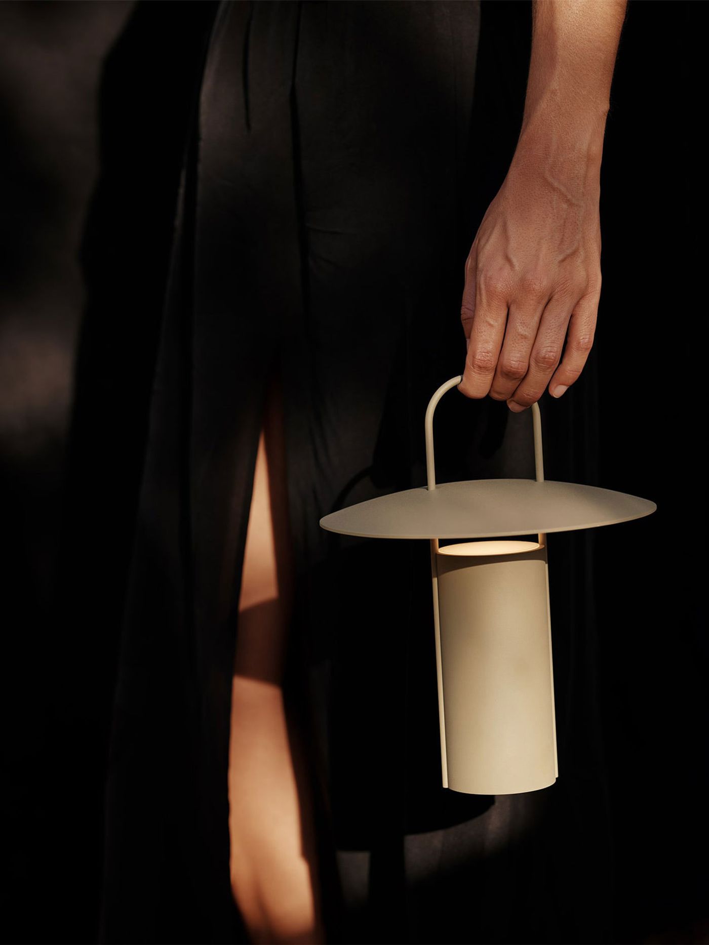 A person in a flowing black dress holds the Ray portable table lamp by Daniel Schofield for Audo; its sleek, sand-colored silhouette and disc-shaped shade stand out against the dramatic shadows. A person in a flowing black dress holds the Ray portable table lamp by Daniel Schofield for Audo; its sleek, sand-colored silhouette and disc-shaped shade stand out against the dramatic shadows.