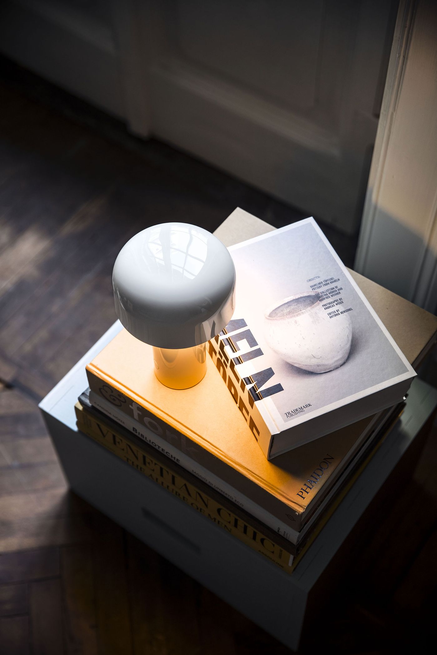 A glossy white Bellhop lamp sits atop a curated stack of design books by a sunlit corner, blending functional light with an editorial interior vignette. A glossy white Bellhop lamp sits atop a curated stack of design books by a sunlit corner, blending functional light with an editorial interior vignette.
