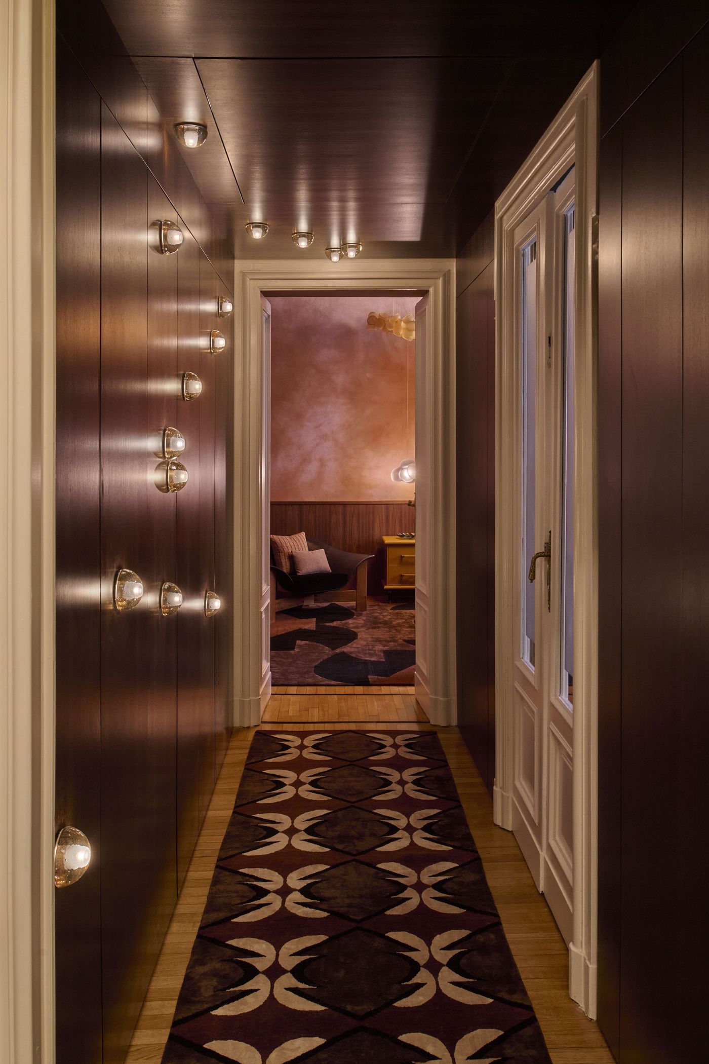 A moody, luxurious corridor lined with dark wood paneling and dotted with spherical wall and ceiling light fixtures, leading to a softly lit room with blush-toned walls, sculptural lighting, and geometric rugs—where mid-century elegance meets contemporary warmth. A moody, luxurious corridor lined with dark wood paneling and dotted with spherical wall and ceiling light fixtures, leading to a softly lit room with blush-toned walls, sculptural lighting, and geometric rugs—where mid-century elegance meets contemporary warmth.