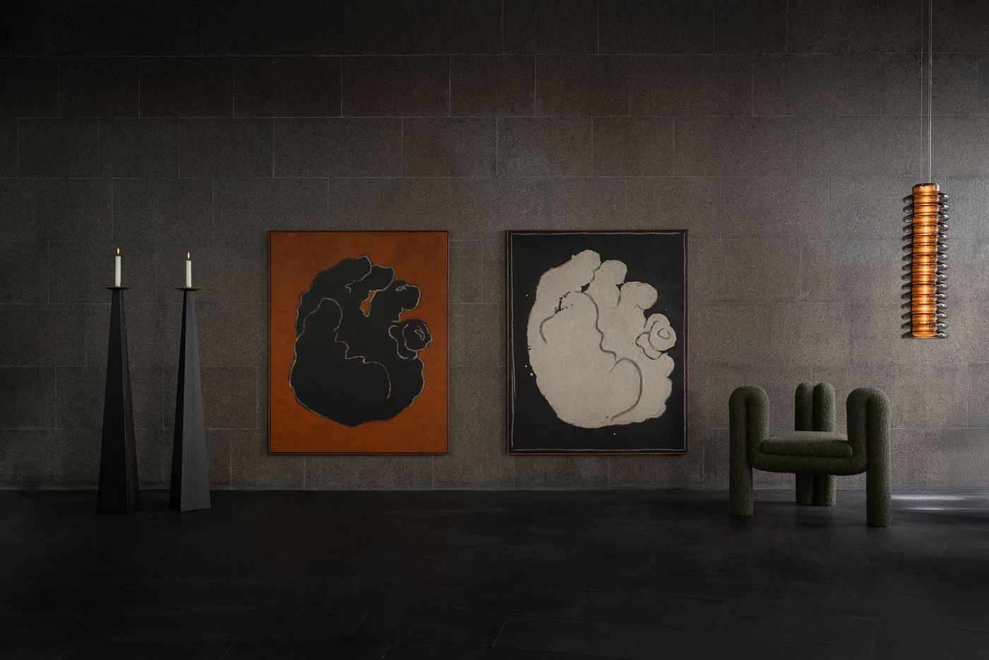 Two monochrome paintings of abstract hands hang on a stone wall, flanked by black candleholders and a green sculptural armchair beneath a cylindrical hanging light.
