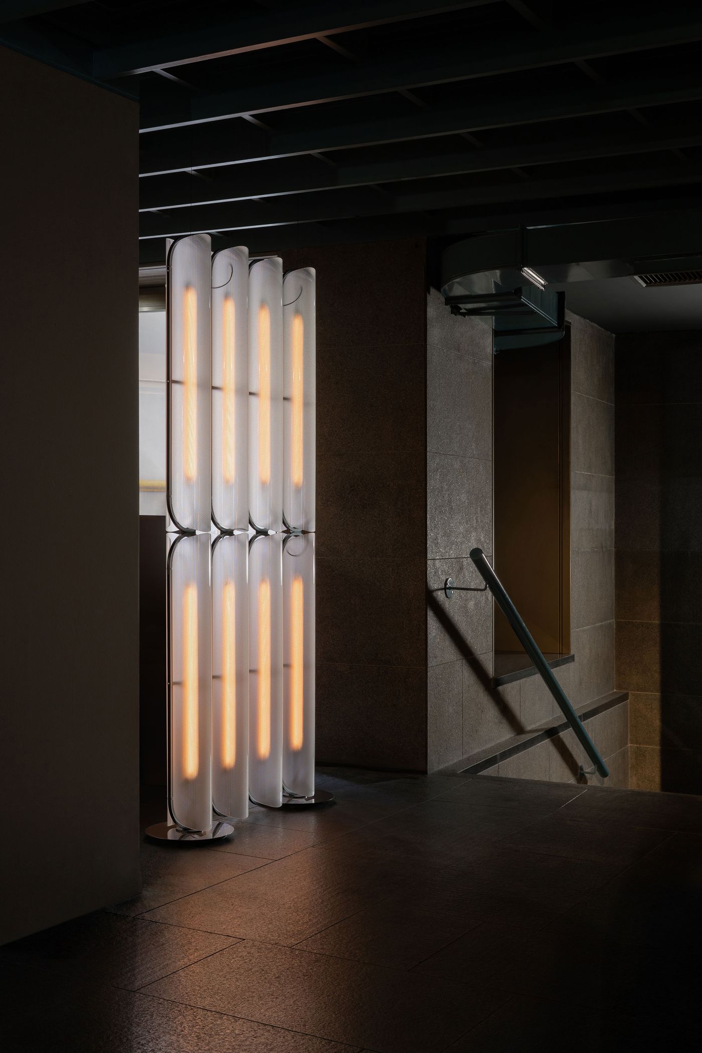 Tall sculptural light fixtures composed of softly glowing vertical glass tubes stand elegantly in a dimly lit corner with stone flooring and walls.