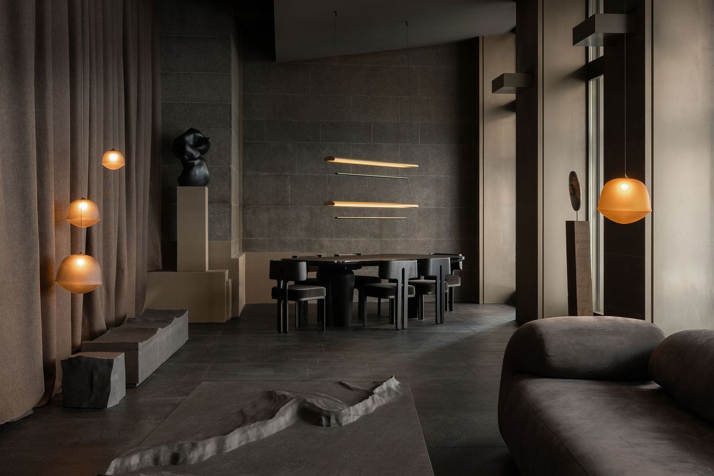 A moody dining room with layered lighting, minimal sculptural furniture, earth-toned curtains, and textured stone flooring creating a monastic-modern ambiance.