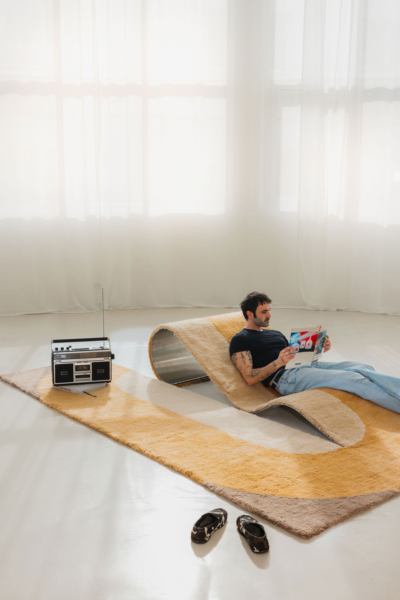 A man lounges on a looped rug-chair hybrid with a retro radio nearby—soft nostalgia meets sculptural comfort. A man lounges on a looped rug-chair hybrid with a retro radio nearby—soft nostalgia meets sculptural comfort.