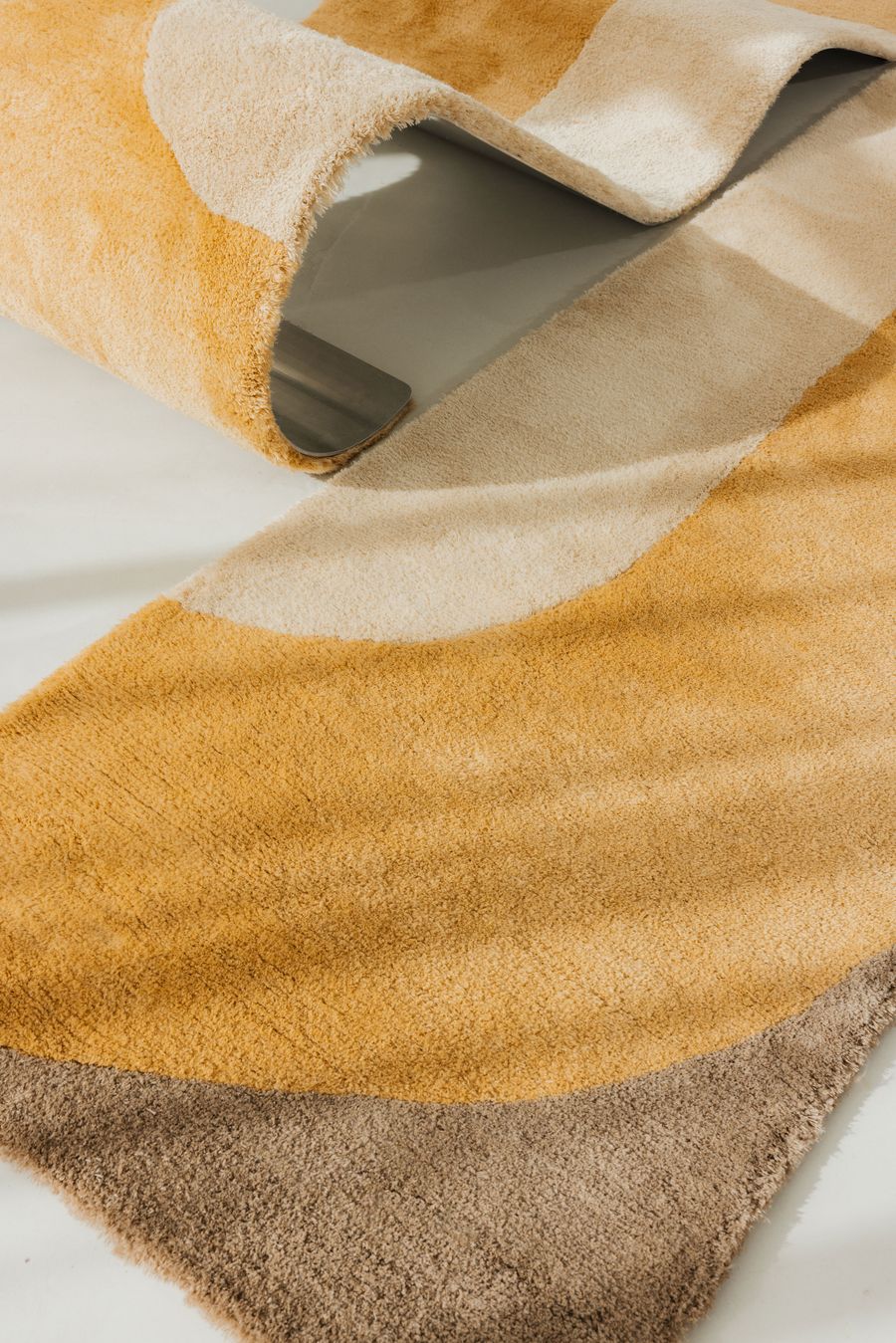 Close-up of a wavy yellow and beige sculptural rug that integrates metal to hold its form. Close-up of a wavy yellow and beige sculptural rug that integrates metal to hold its form.