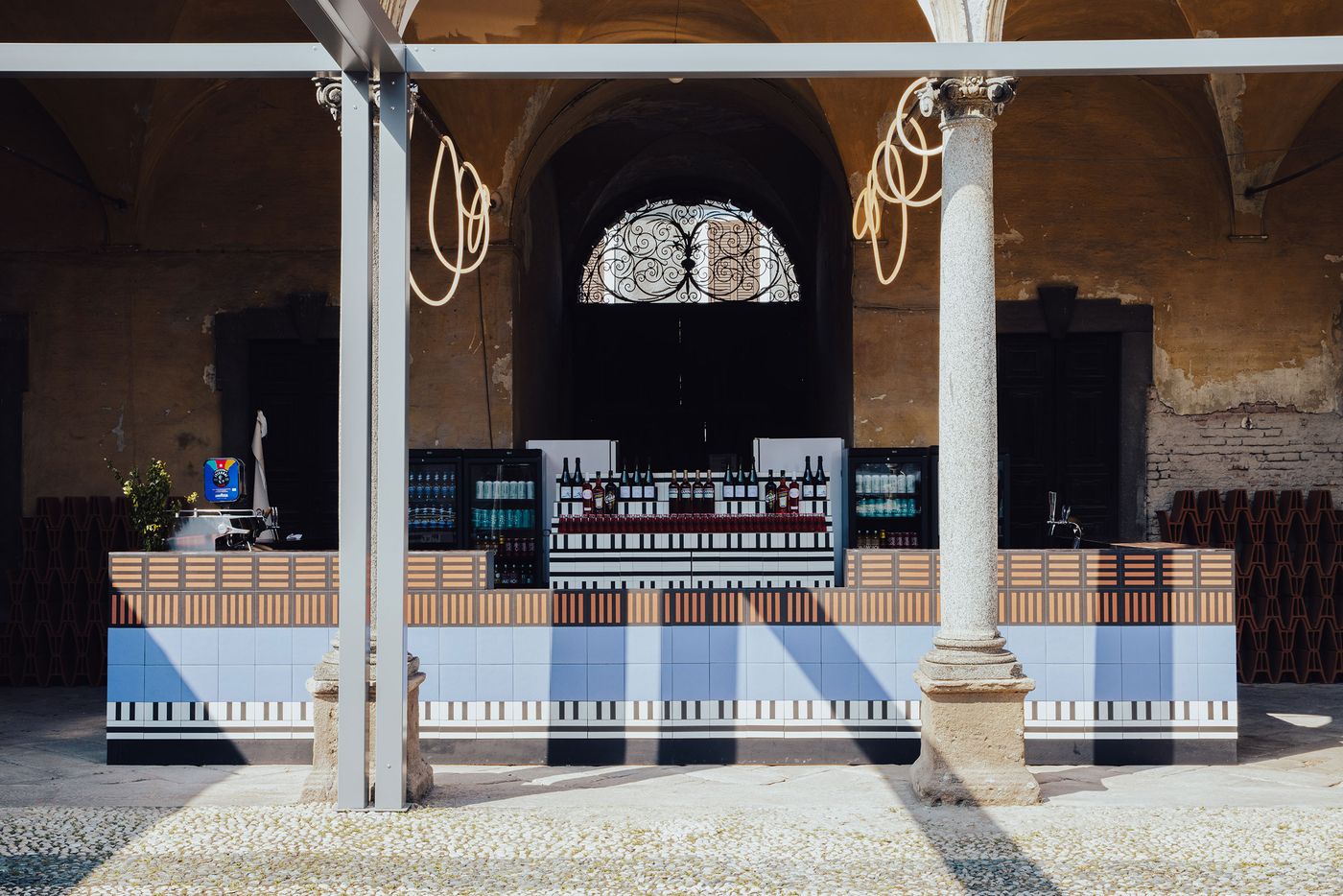 A vibrant display of bottles behind a checker-patterned bar, set within a sunlit portico—drinks served with a dose of design.