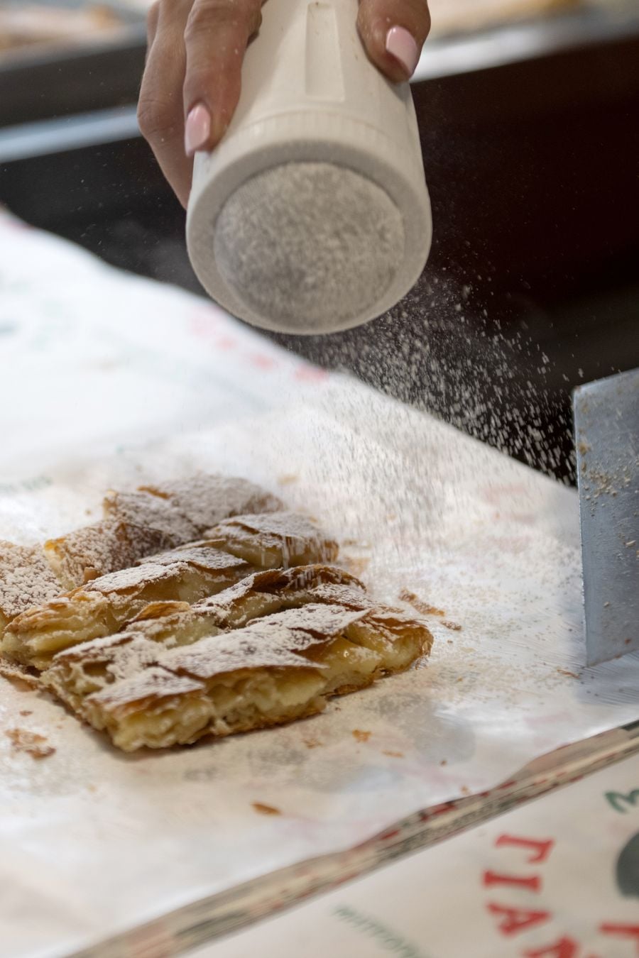 Close-up of freshly baked Greek bougatsa at Bougatsa Giannis in Thessaloniki, its golden, flaky pastry cut into pieces and generously dusted with powdered sugar. A hand shakes more sugar over the pastry, capturing the preparation of one of the city’s most beloved culinary specialities.
