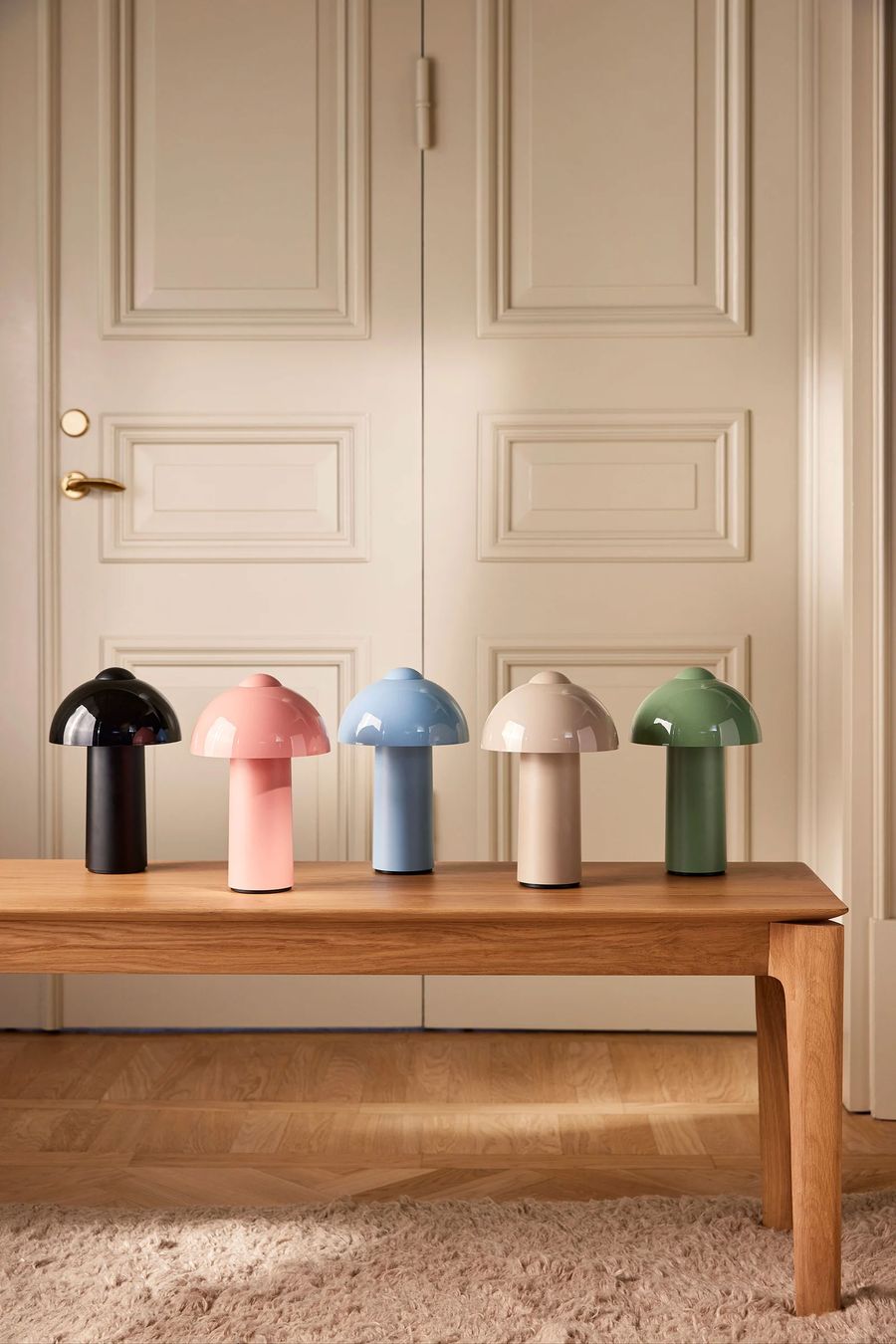 Five mushroom-shaped table lamps in glossy finishes—black, pink, blue, beige, and green—are neatly arranged on a wooden bench in front of double cream-colored paneled doors, with a light wood floor and a soft beige rug beneath. Five mushroom-shaped table lamps in glossy finishes—black, pink, blue, beige, and green—are neatly arranged on a wooden bench in front of double cream-colored paneled doors, with a light wood floor and a soft beige rug beneath.
