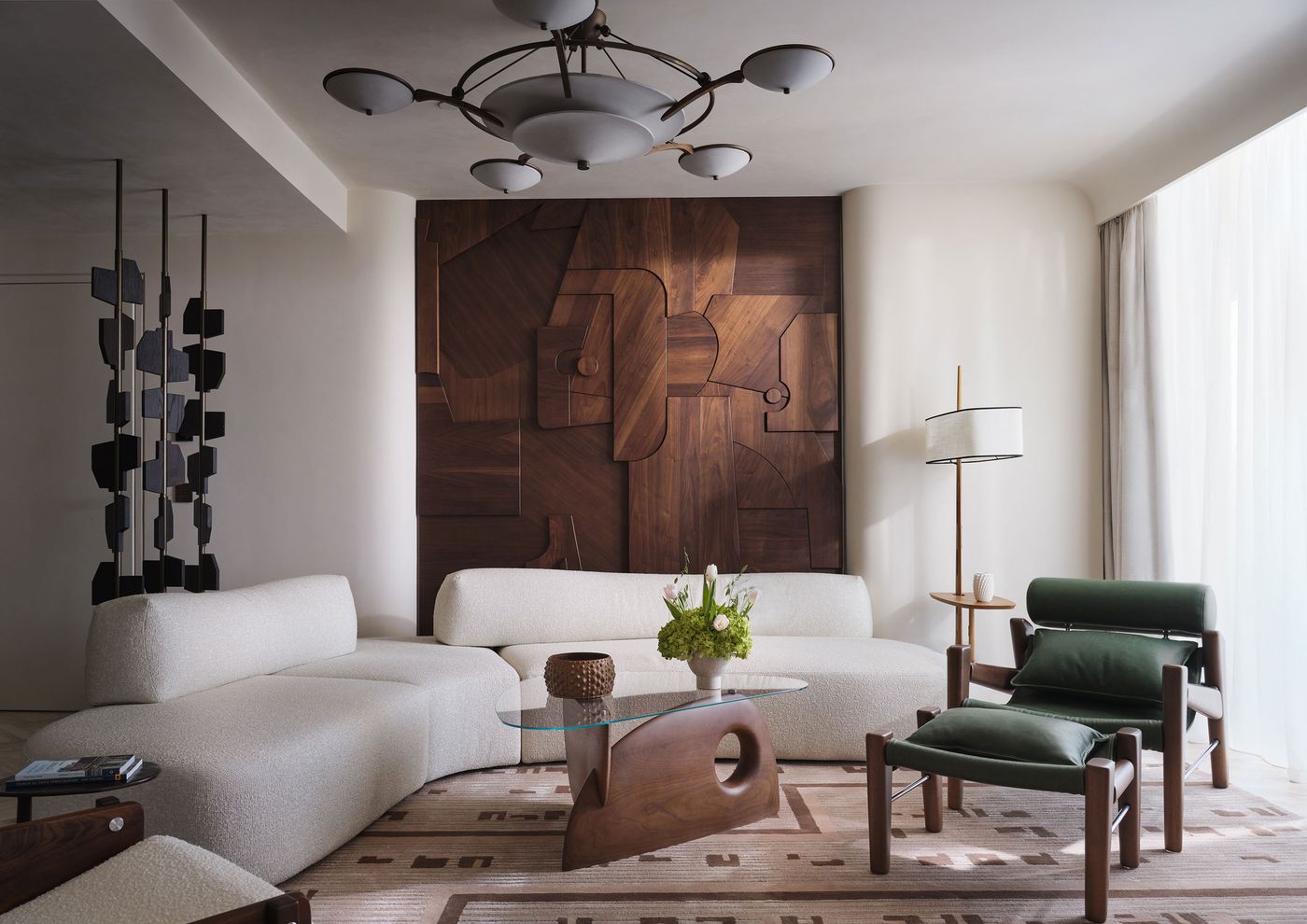 An elegant suite lounge at Conrad Athens combines a sweeping white bouclé sectional sofa, deep-green upholstered armchair and sculptural timber-and-glass coffee table. A large geometric walnut wall relief anchors the composition, complemented by a modernist ceiling fixture, patterned rug and black sculptural room divider. Soft daylight highlights the interplay of rich wood, tactile fabrics and contemporary forms.