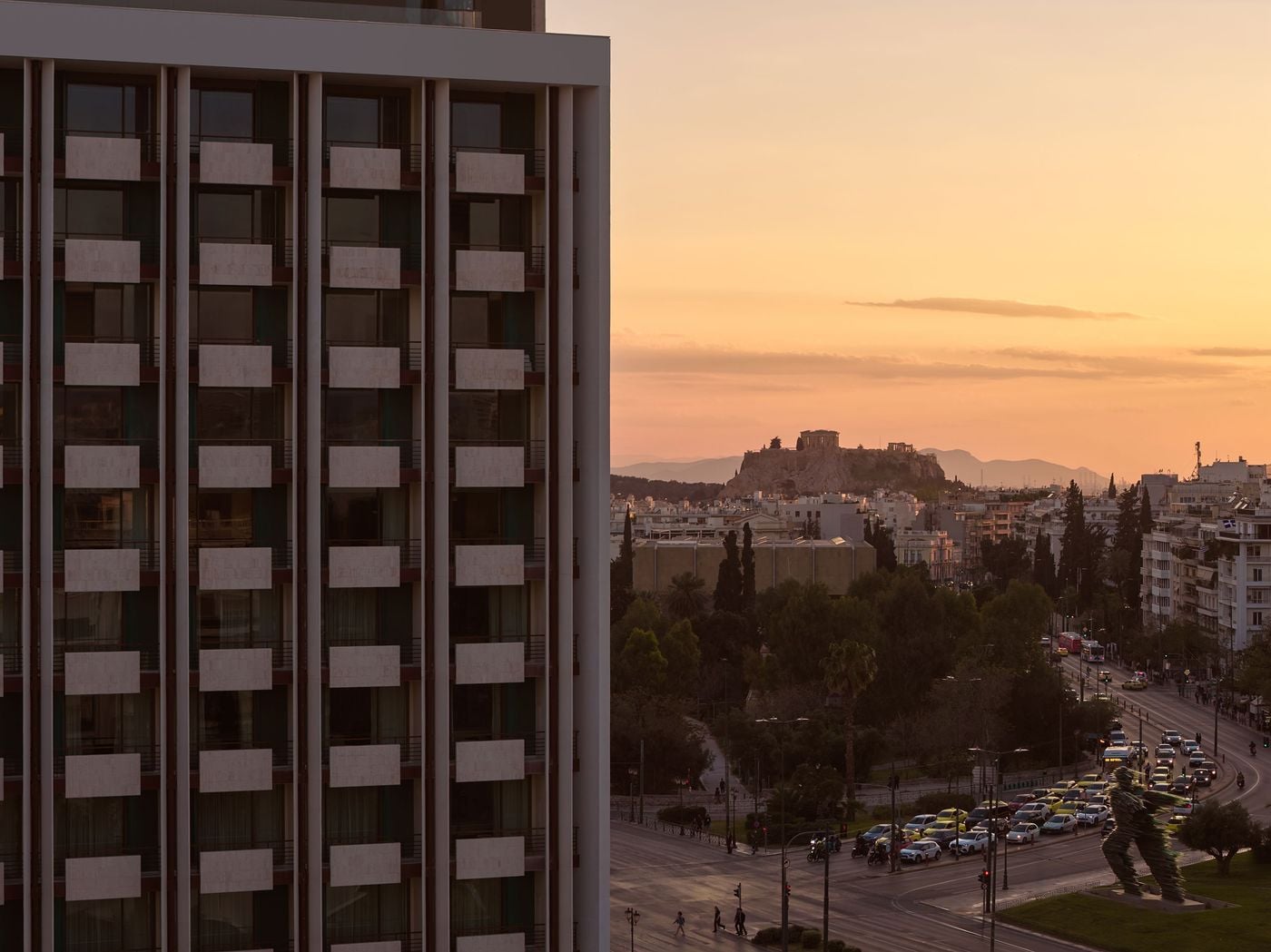 A wider urban view situates the building within Athens, with the Acropolis rising in the distance. The contrast between the hotel’s ordered façade and the layered cityscape underscores its presence within the evolving urban fabric.