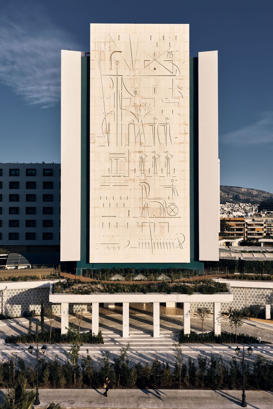 The monumental façade of Conrad Athens at The Ilisian features a towering pale stone mural engraved with abstract linear figures and geometric motifs. Flanked by stark white vertical volumes, the modernist building rises above a landscaped colonnaded entrance, its architectural symmetry and sculptural artwork standing in contrast to the surrounding Athenian skyline.