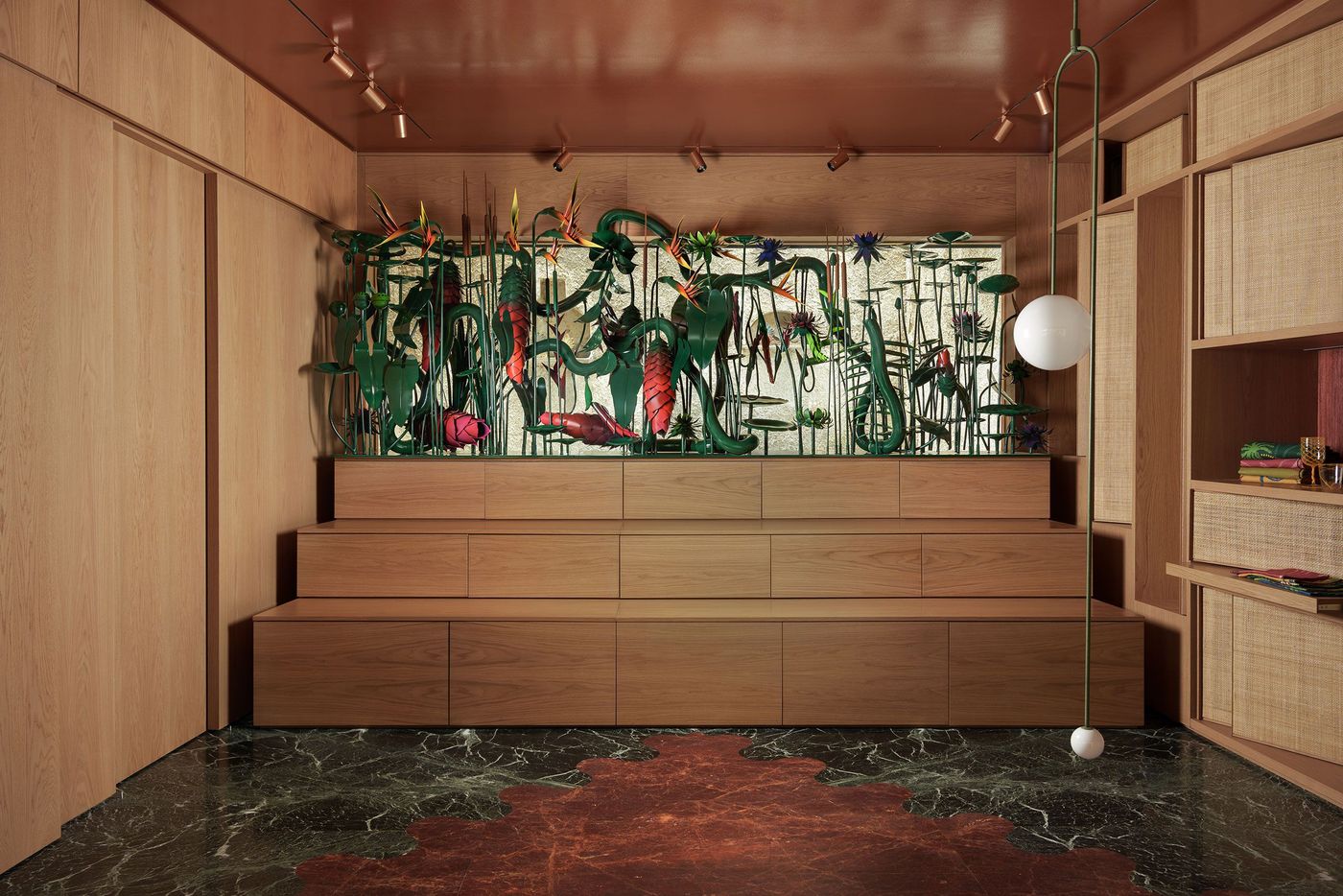 A striking installation of sculptural metal flowers and tropical foliage dominates the Crini & Sophia showroom in Athens, framed by warm timber panelling and tiered wooden cabinetry. The elaborate botanical composition, featuring green stems and vivid pink and orange blooms, contrasts with the burgundy ceiling and graphic green-and-red marble flooring, creating an eclectic, theatrical atmosphere.