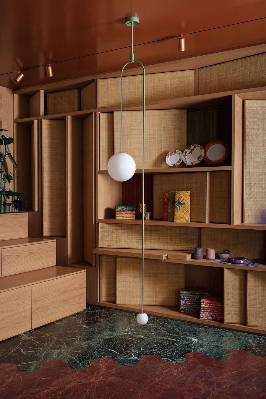 The richly textured interior of Crini & Sophia's Athens showroom features bespoke timber shelving with woven panels displaying colourful ceramics, patterned fabrics and decorative objects. A slender green metal pendant with spherical white glass lights hangs against a terracotta ceiling, while contrasting green and burgundy marble flooring adds a graphic flourish to the warm, eclectic composition.