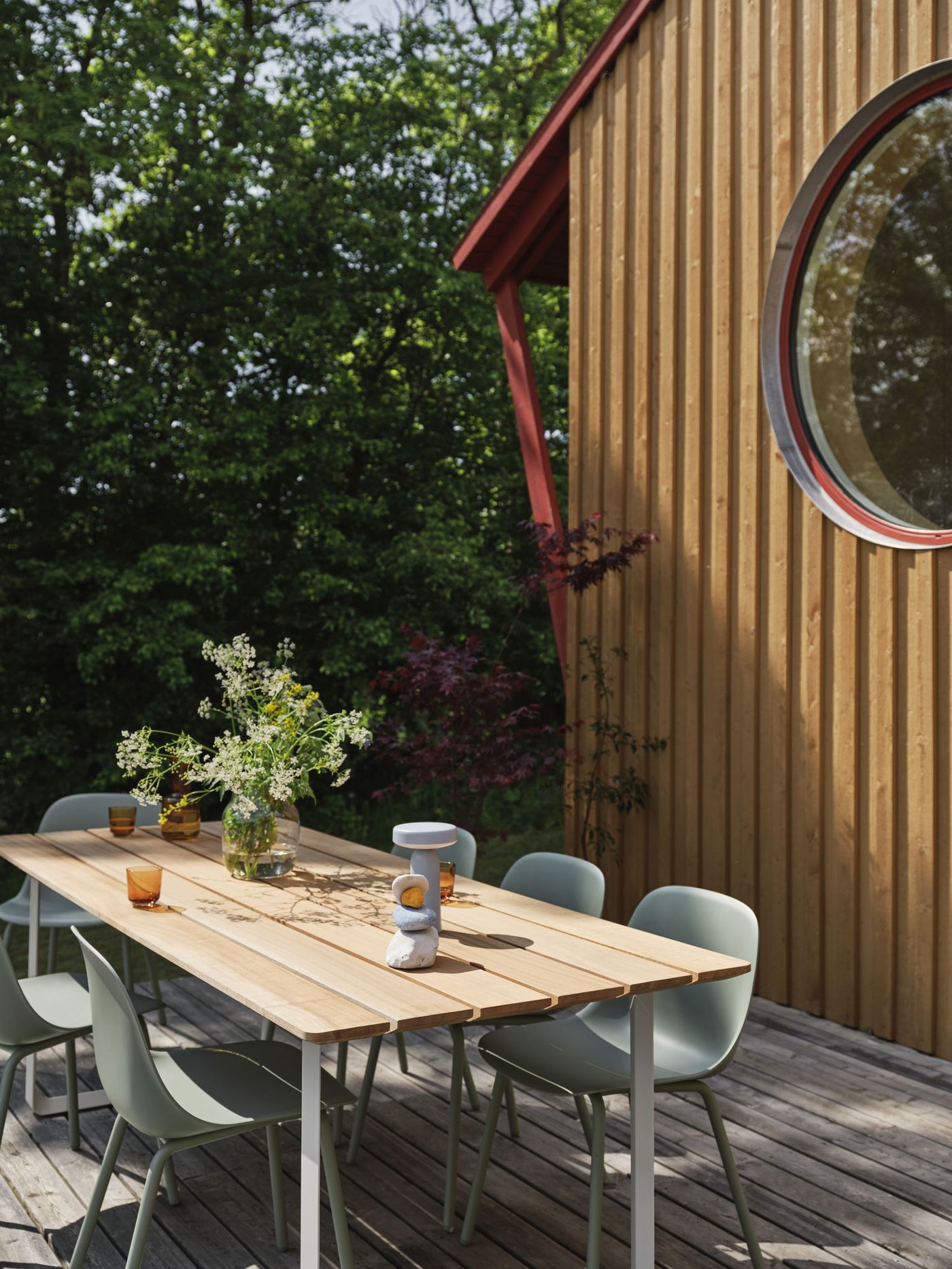EASE Portable Lamp sits on a wooden outdoor table alongside sculptural decor, amber glasses, and a wildflower bouquet, framed by soft green chairs and a timber-clad house with a circular window.