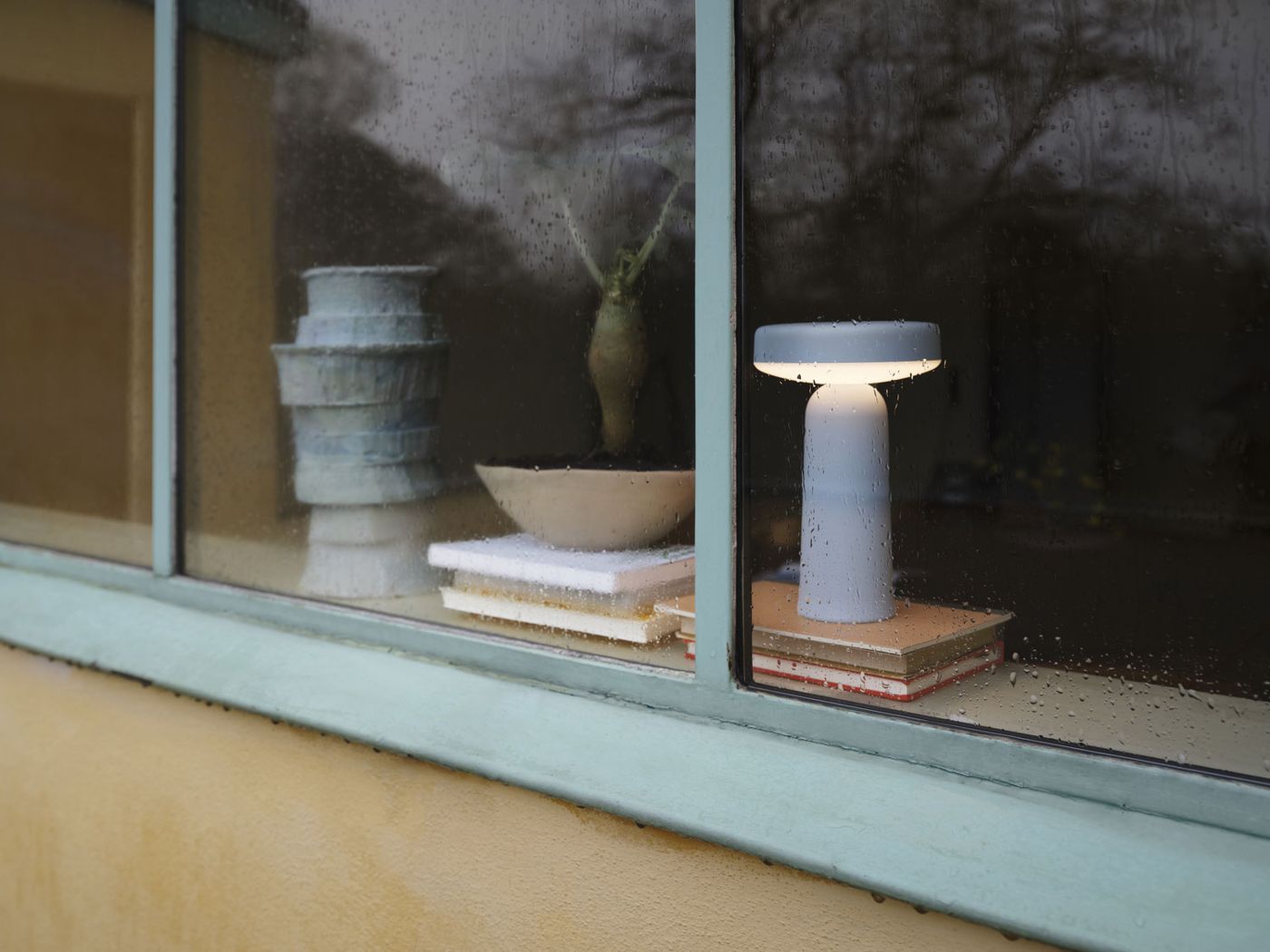 EASE blue portable lamp softly glowing indoors on a windowsill, seen through a rain-speckled window, resting atop stacked books beside ceramic objects.