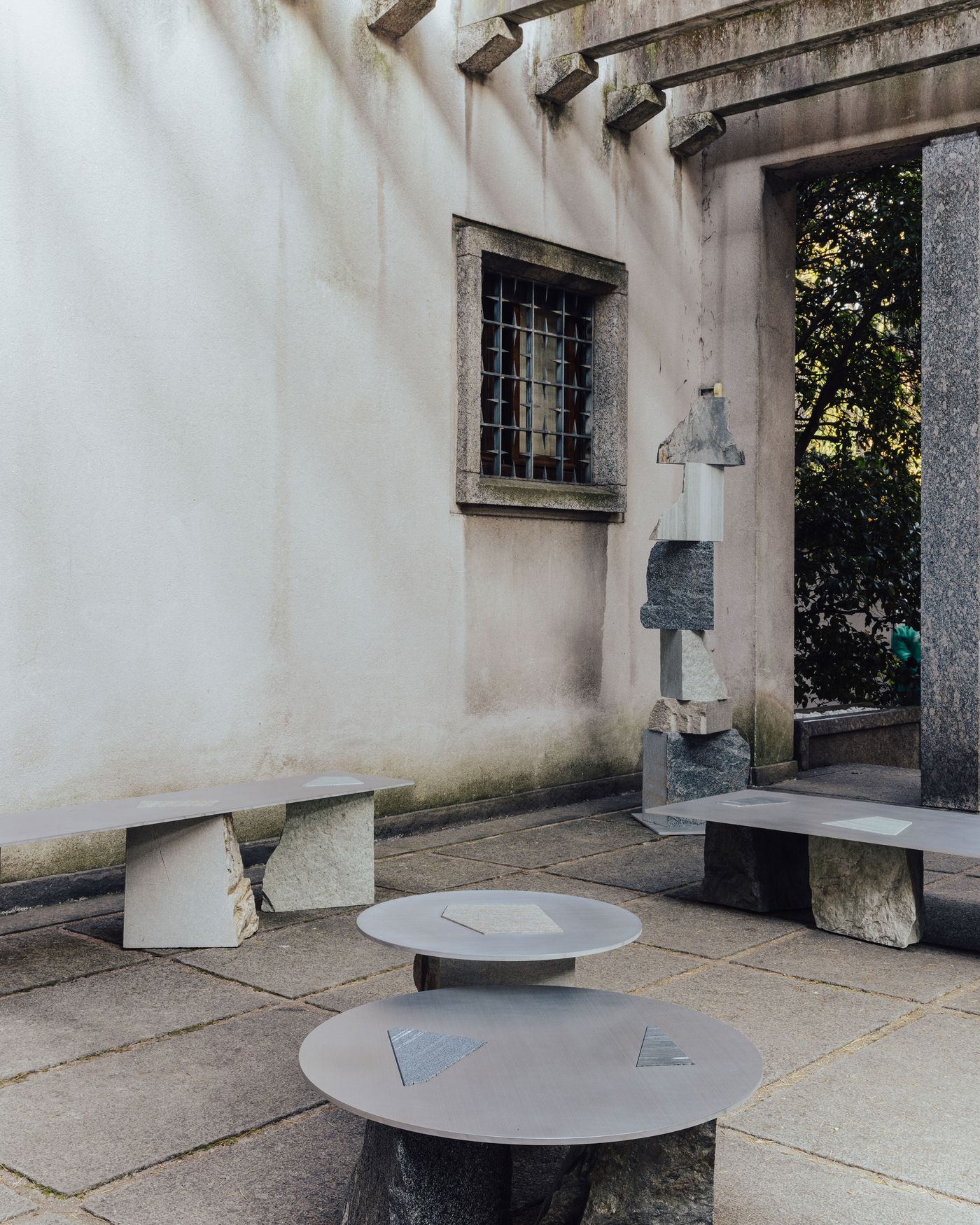 Minimalist stone and metal furniture pieces by Estudio Material at Villa Borsani, framed by aged stone walls, a gridded window, and dappled sunlight. Minimalist stone and metal furniture pieces by Estudio Material at Villa Borsani, framed by aged stone walls, a gridded window, and dappled sunlight.