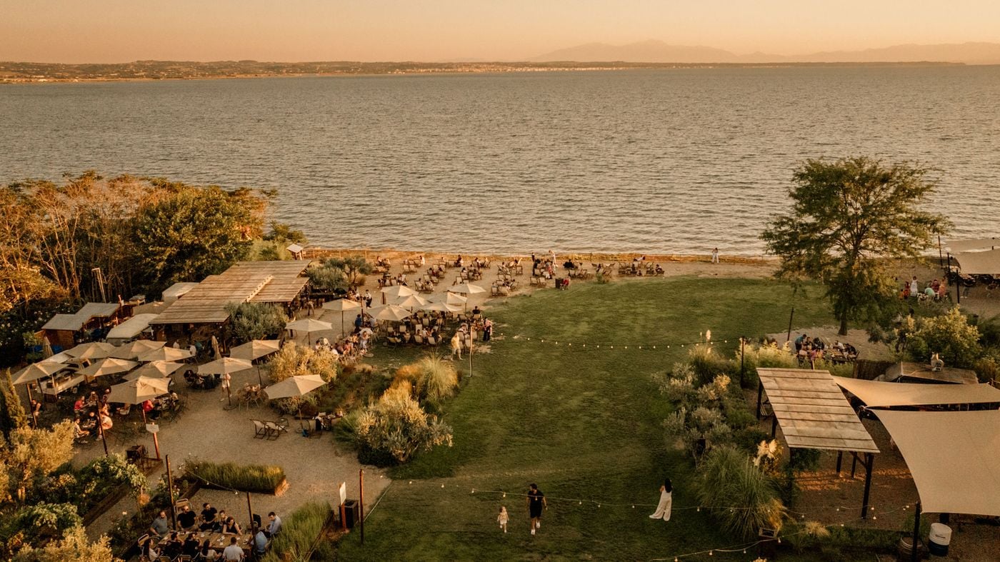 Elevated view of Ergon Agora East in Thessaloniki at sunset, where a sprawling waterfront dining destination unfolds around an expansive green lawn. Timber pergolas, cream parasols, planted borders and outdoor seating areas accommodate guests enjoying the evening, with the sea and distant mountains forming a tranquil backdrop.