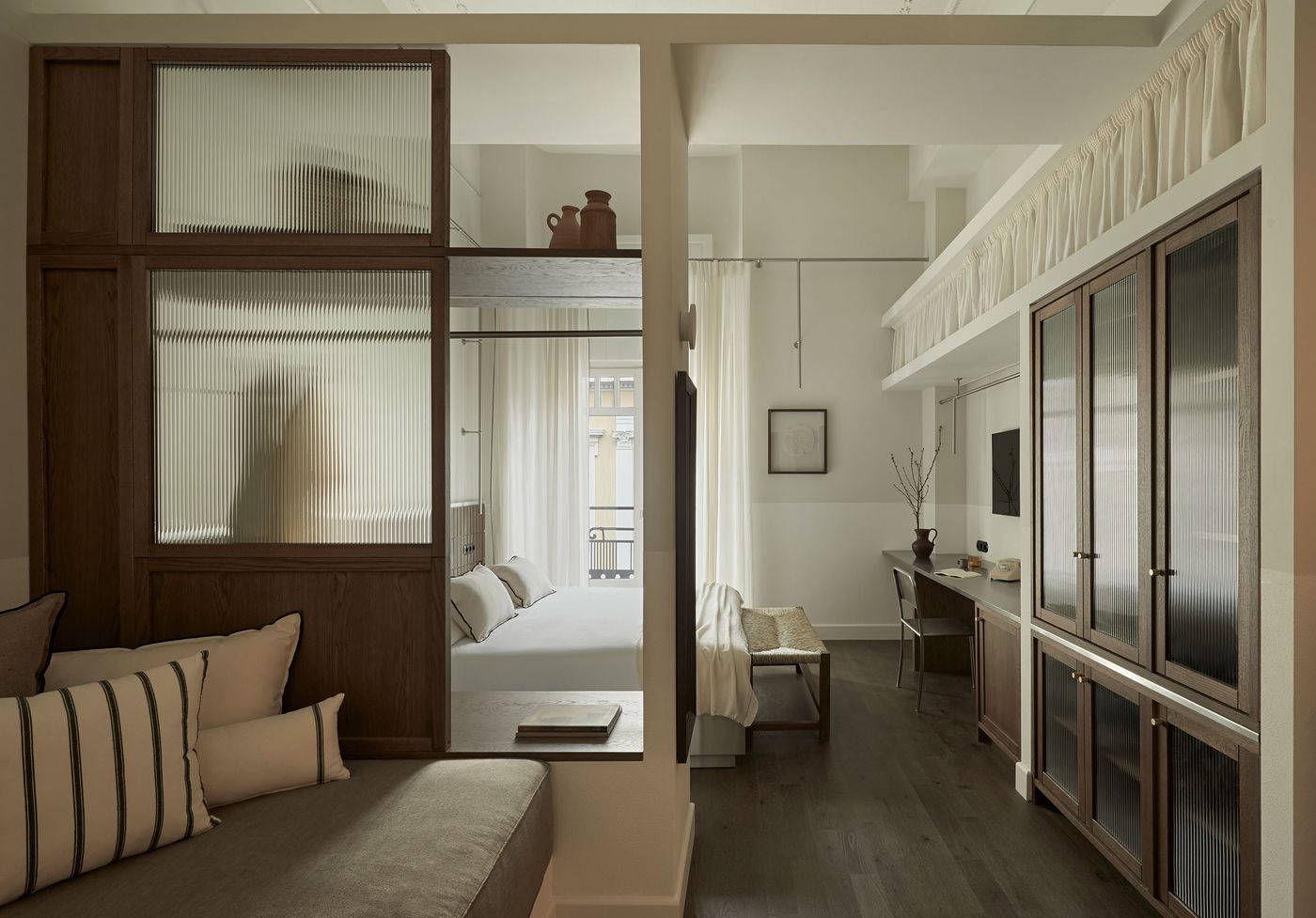 A contemporary guest room at Ergon Bakehouse in Athens combines warm walnut-toned joinery, ribbed glass partitions and muted neutral fabrics. A built-in sofa occupies the foreground, while timber-framed screens partially separate the sleeping and working areas. Dark wooden flooring, minimalist furniture and softly diffused daylight create an atmosphere of restrained, Japanese-inspired simplicity.