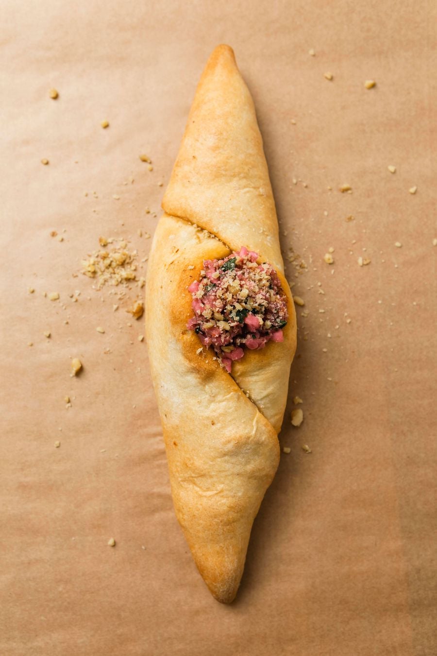 Overhead close-up of a golden-baked peinirli from Feyrouz in Thessaloniki, its elongated, boat-shaped dough folded around a savoury minced-meat filling garnished with herbs and crushed nuts. Presented on brown parchment paper, the pastry showcases the bakery’s Middle Eastern-inspired culinary repertoire.