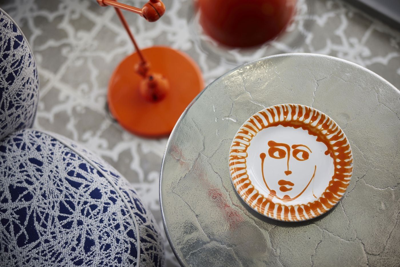 A close-up of a hand-painted ceramic plate featuring an abstract ochre-coloured face, resting on a textured silver tabletop at Fresh Hotel in Athens. Blue-and-white patterned upholstery and vivid orange lighting fixtures surround the composition, capturing the hotel's eclectic mix of artisanal decorative objects, graphic motifs and playful Mediterranean colours.