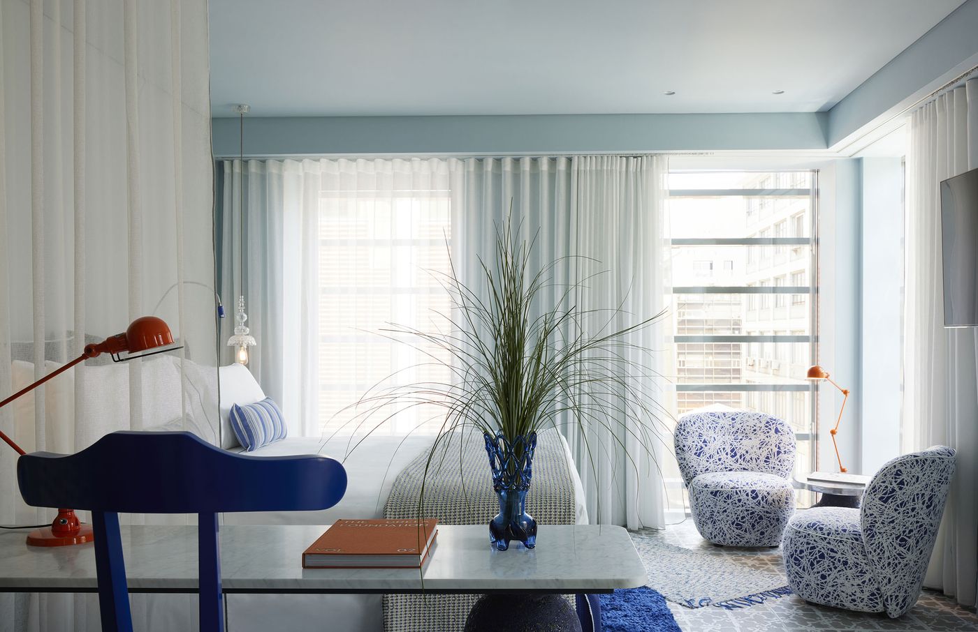 A light-filled guest room at Fresh Hotel in Athens showcases designer Paola Navone’s playful approach to colour and texture. Cobalt-blue furnishings, patterned upholstered armchairs, vivid orange task lamps and a sculptural blue glass vase punctuate the predominantly white interior. Sheer curtains, patterned flooring and expansive windows create an airy, contemporary atmosphere with Mediterranean flair.