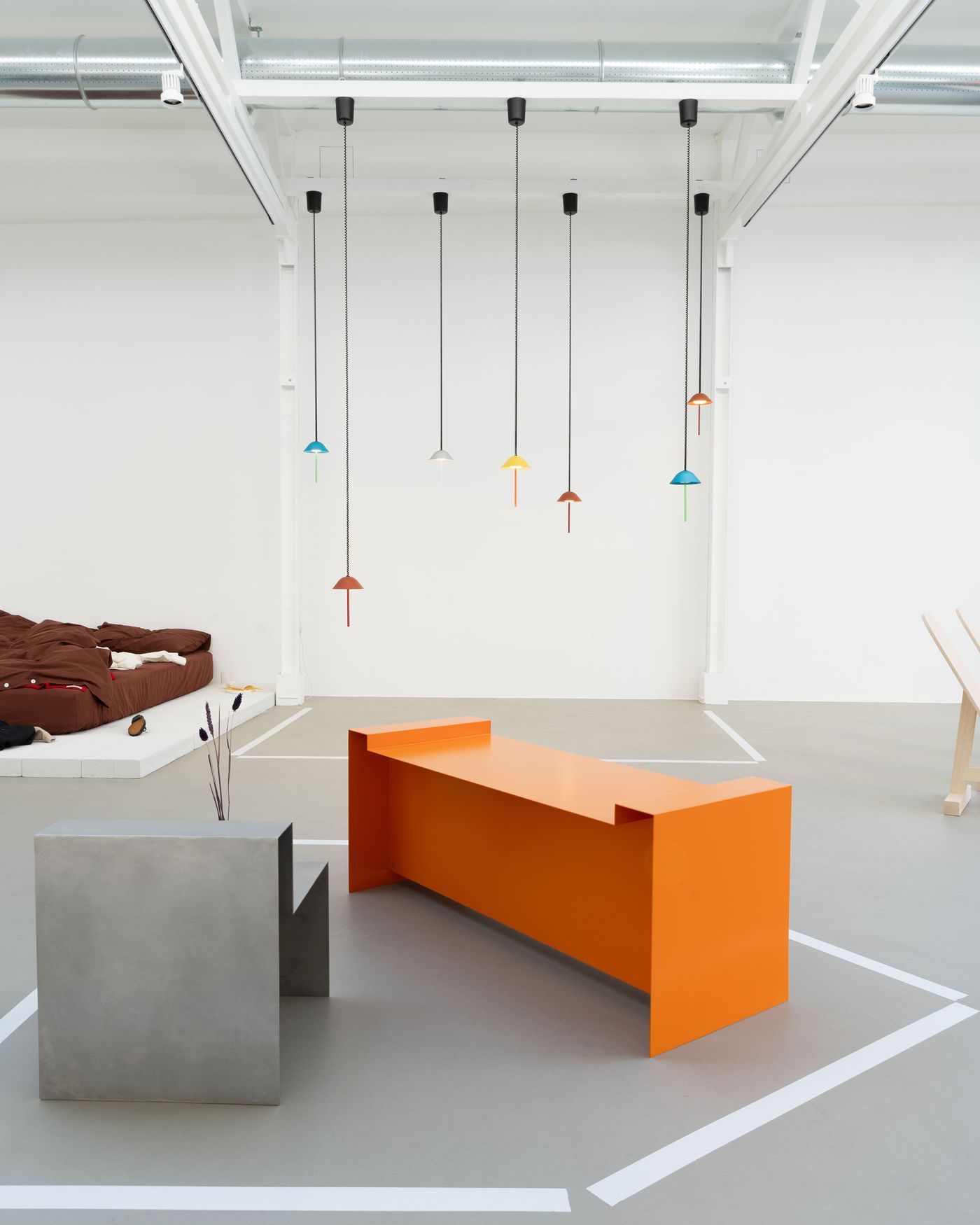 A minimal showroom with vibrant pendant lights, a bold orange bench, and a concrete cube chair under an airy white ceiling.