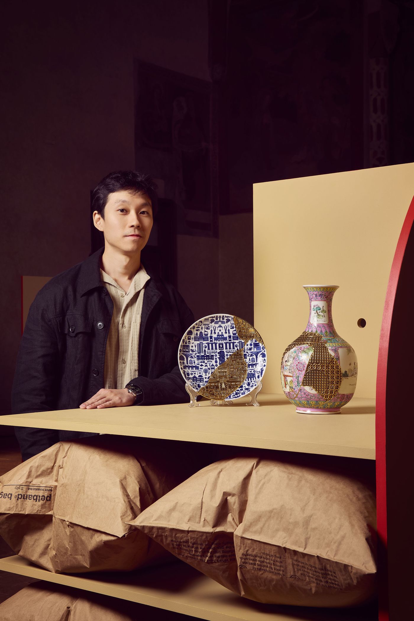 A man posing next to a ceramic plate and a vase, both decorated with intricate golden repairs, set on a minimalist mustard-colored display.