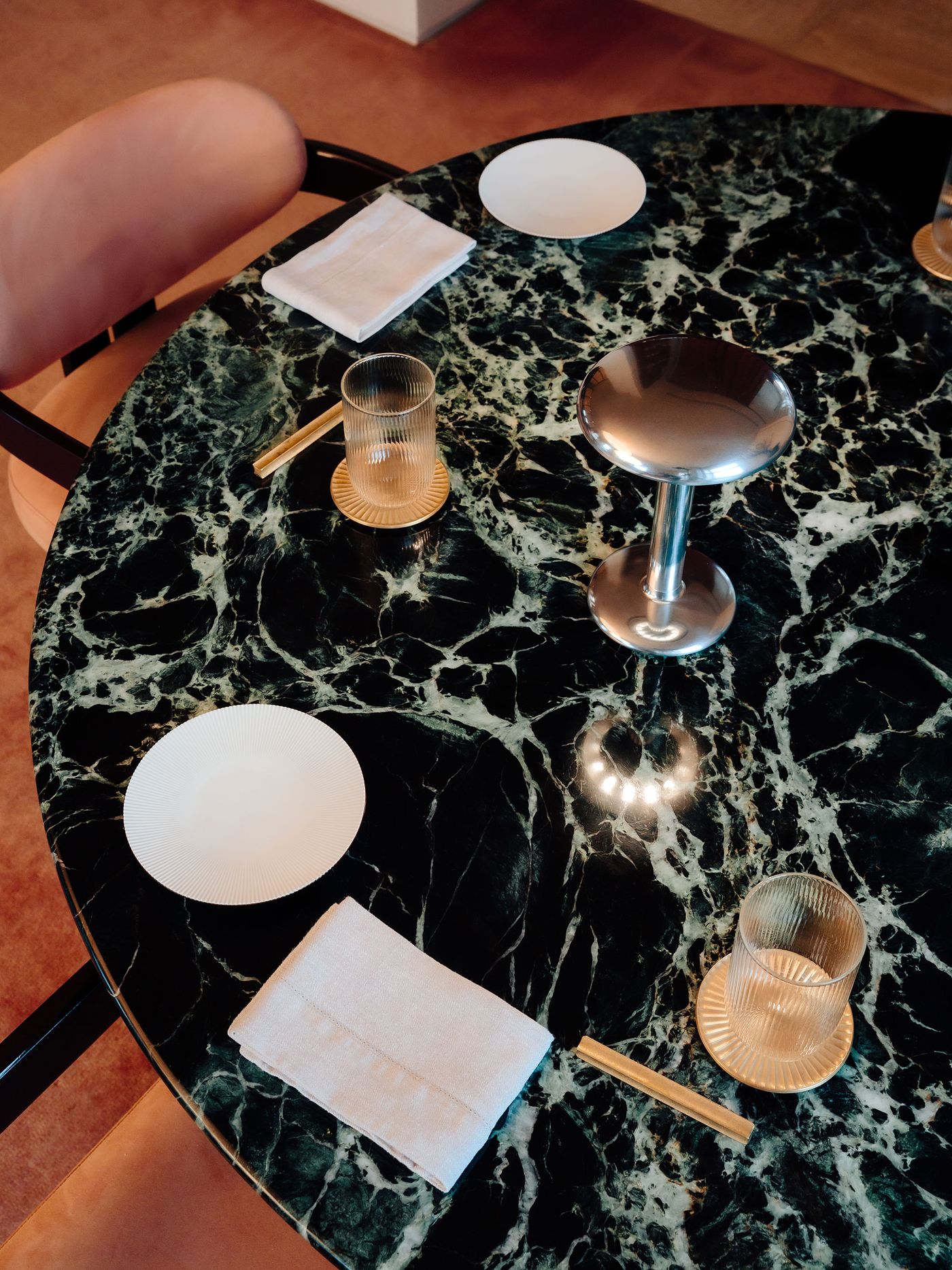 A polished metal Gustave lamp sits centered on a round green marble table surrounded by ribbed glassware, gold-rimmed plates, and soft pink velvet chairs. A polished metal Gustave lamp sits centered on a round green marble table surrounded by ribbed glassware, gold-rimmed plates, and soft pink velvet chairs.
