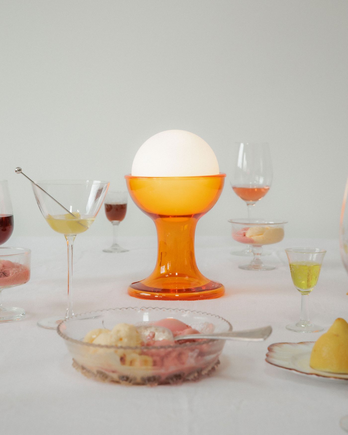 An orange glass Gelato table lamp with a glowing white orb top, styled as a centrepiece on a dessert table with ice cream, glassware, and fruit under soft daylight. An orange glass Gelato table lamp with a glowing white orb top, styled as a centrepiece on a dessert table with ice cream, glassware, and fruit under soft daylight.