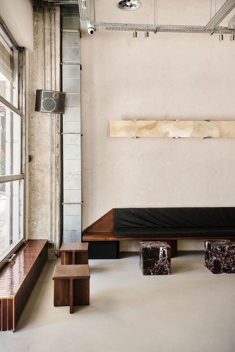 A minimalist, industrial-inspired interior at He-art in Thessaloniki, featuring a long timber bench with black cushions, burgundy marble cube tables and compact wooden stools. Exposed ventilation ducts, raw concrete surfaces and a pale plaster wall contrast with the warmth of the timber furnishings and natural daylight streaming through large windows.