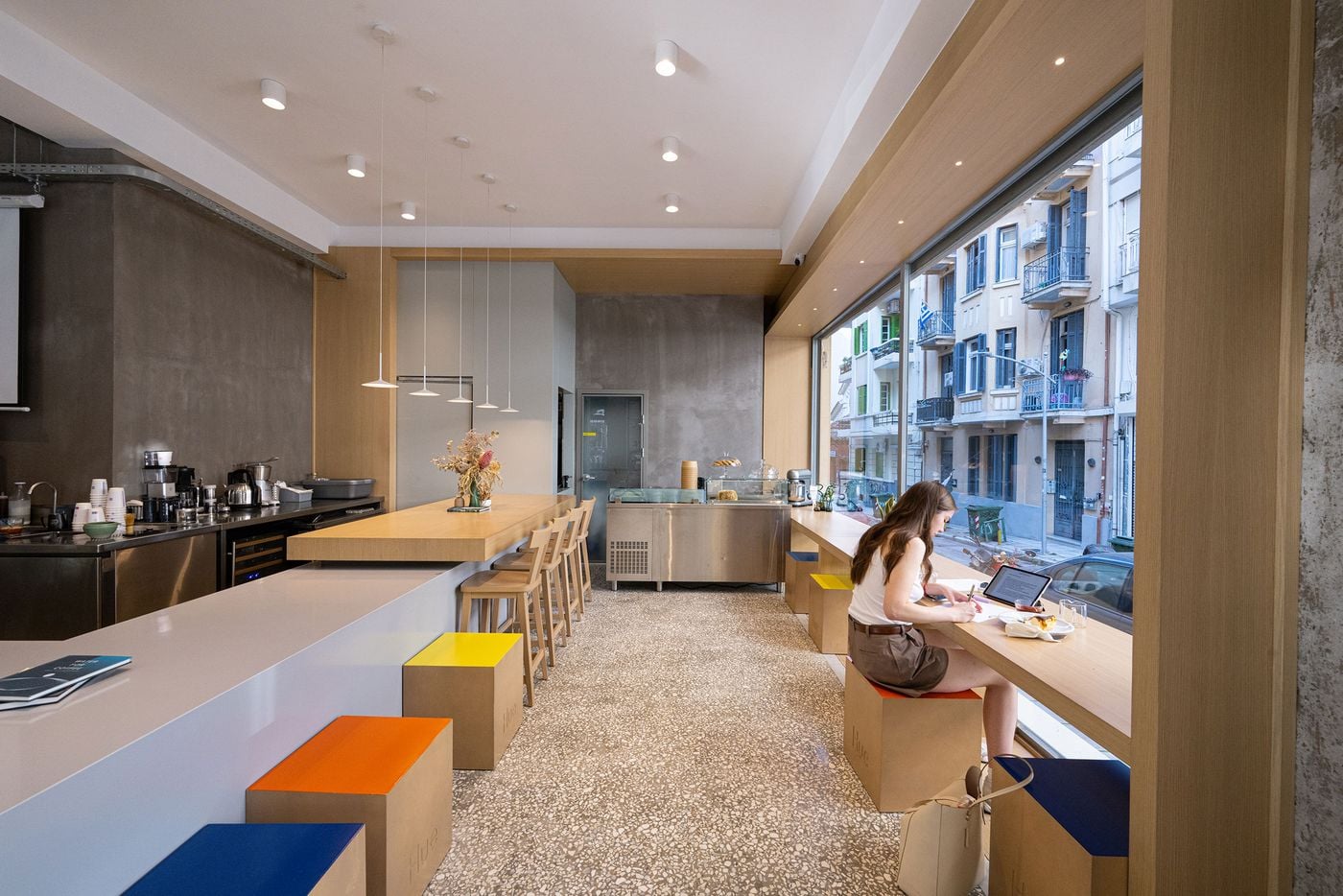The contemporary interior of Hue Roastery in Thessaloniki combines terrazzo flooring, raw concrete walls, pale timber counters and stainless-steel equipment. A long communal counter with wooden stools anchors the minimalist space, while colourful cube seats punctuate the otherwise neutral palette. A customer works beside the expansive street-facing window.