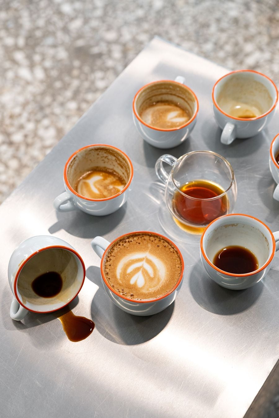 An assortment of espresso cups and a glass coffee vessel arranged across a brushed stainless-steel tabletop at Hue Roastery in Thessaloniki. White ceramic cups with vivid orange rims contain espresso and milk-based coffees, including delicate latte art, while bright sunlight casts crisp shadows across the reflective surface.
