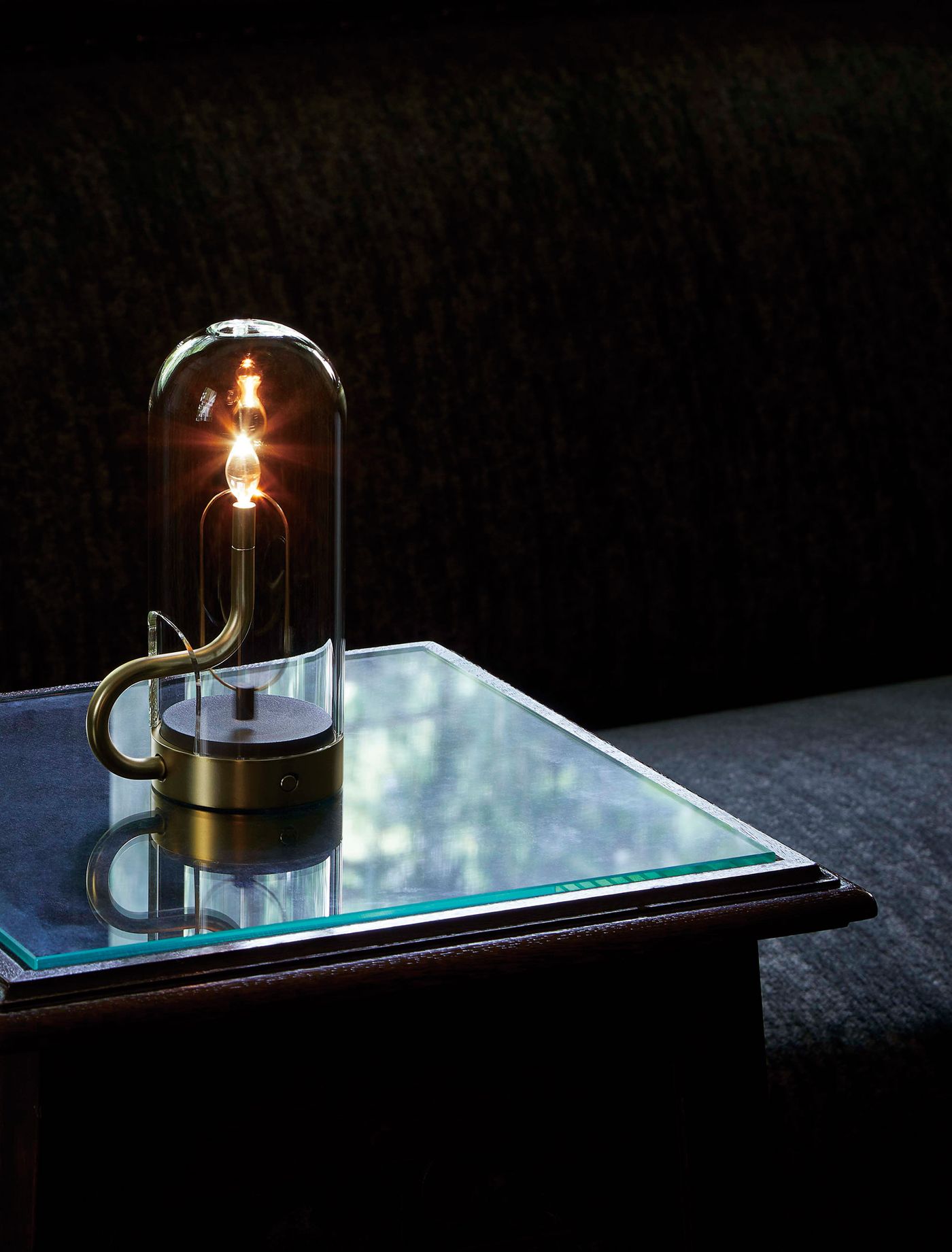 A modern gold and glass table lamp with a glowing filament, encased in a clear dome-shaped glass cover, sits on a reflective glass-topped side table against a dark upholstered background.