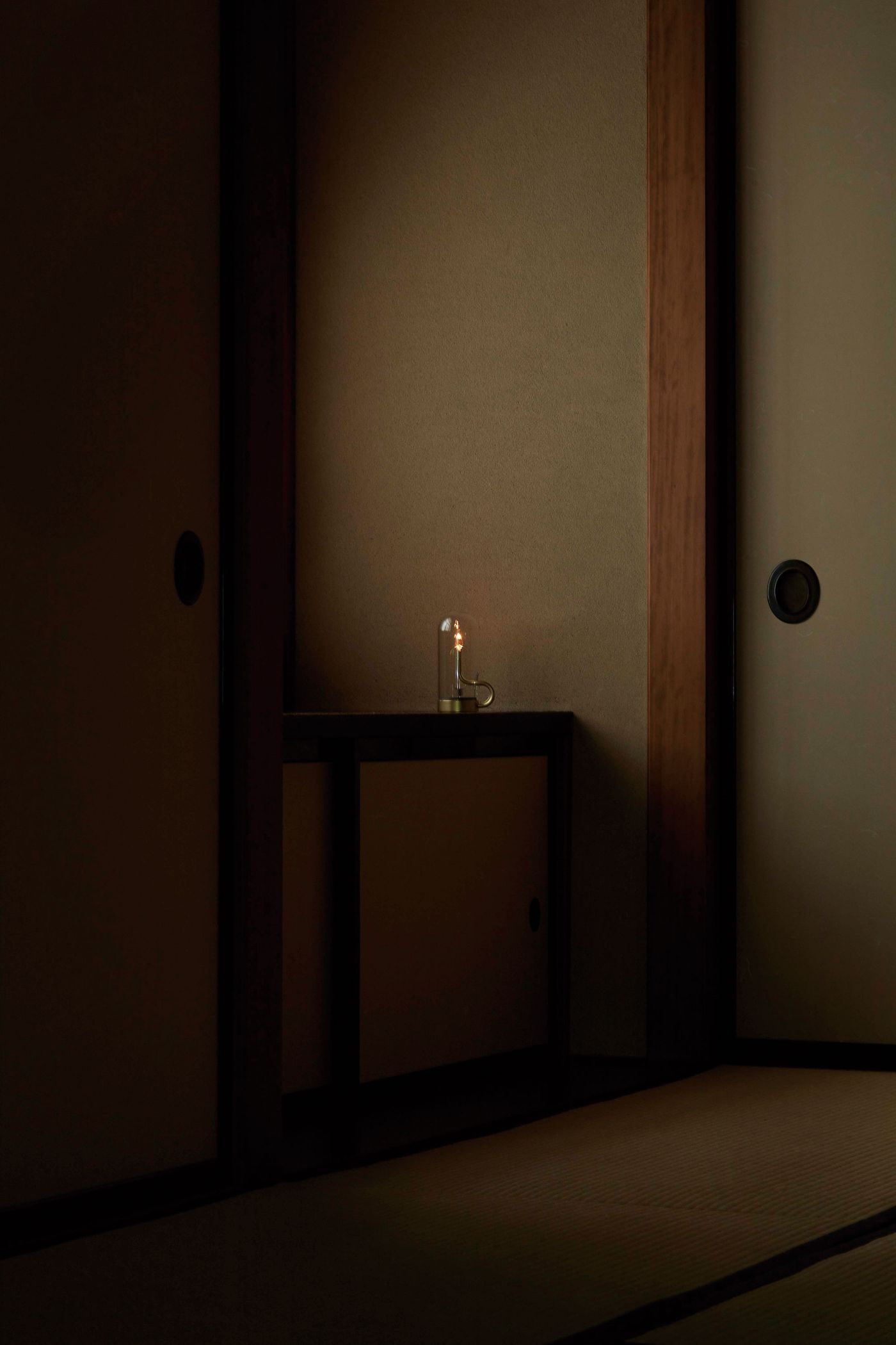 A minimal, serene Japanese interior featuring a single illuminated lamp with a glass dome and brass base, positioned on a dark wooden ledge against a plain wall. The subdued lighting and simple architectural lines evoke a tranquil, contemplative atmosphere.