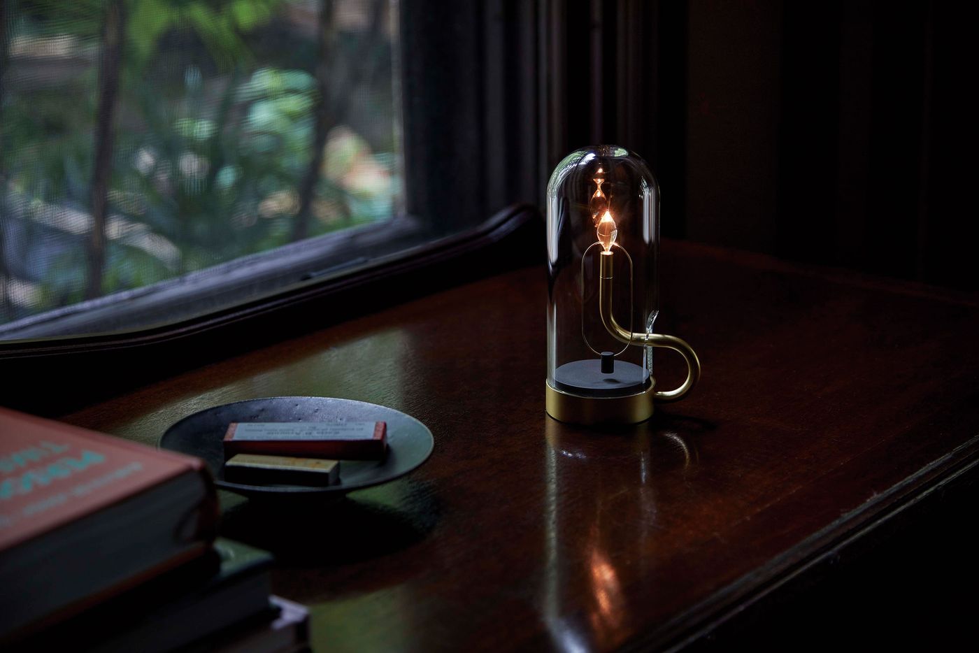 A close-up of a refined table lamp with a glass dome and brass base, glowing warmly on a polished wooden desk near a window. Books and a small tray with matchboxes rest nearby, while greenery is visible through the window in the softly blurred background.