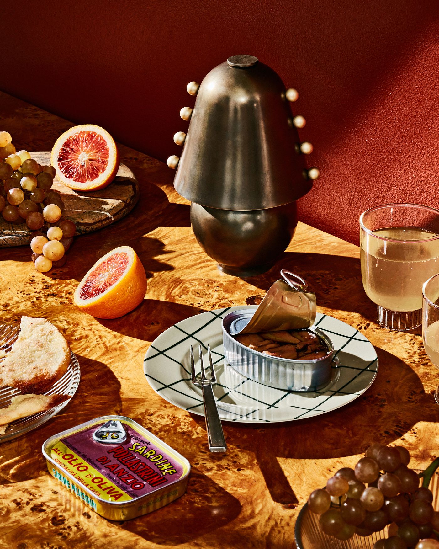 A richly set table with grapes, sliced citrus, tinned sardines, and a pewter-finished Gemma table lamp by In Common With & Sophie Lou Jacobsen as the sculptural centerpiece, softly lit against a deep red wall.