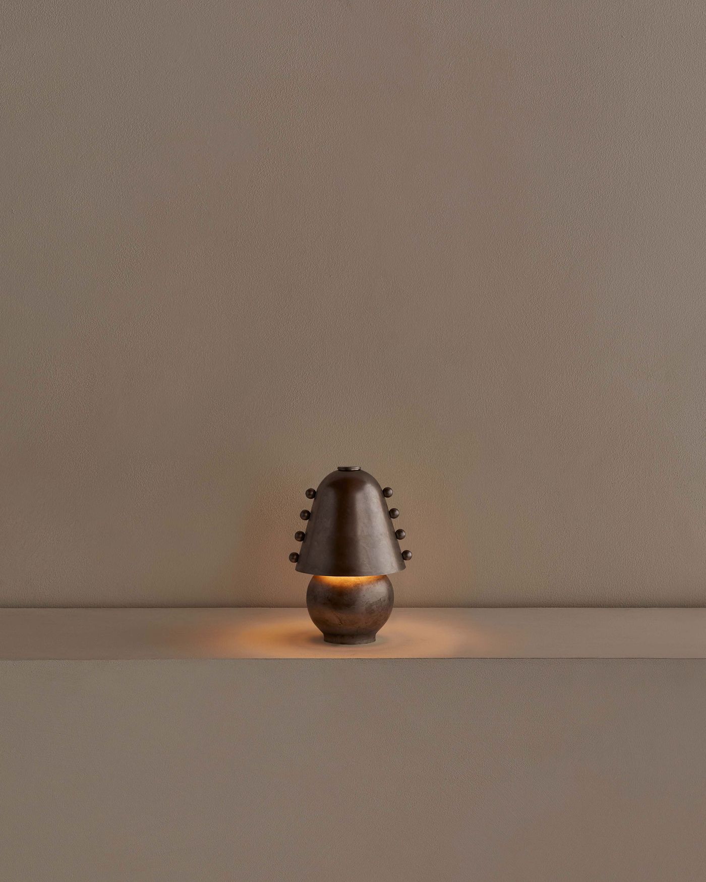 A single pewter-finished Gemma table lamp glows warmly on a minimalist ledge, highlighting its sculptural silhouette against a taupe background.