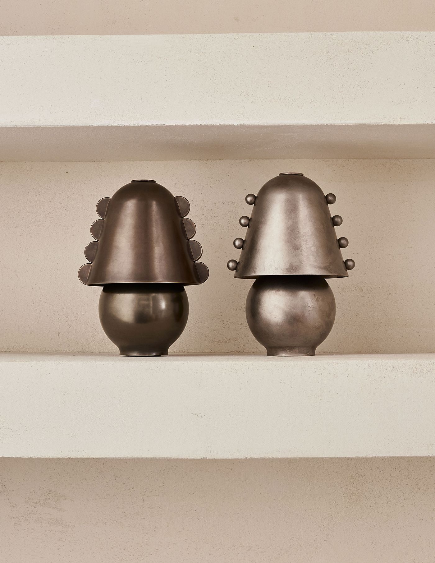 Two Gemma table lamps by In Common With & Sophie Lou Jacobsen sit side-by-side on a plastered shelf—one with disc-like side accents, the other with spherical details—emphasizing their ornamental asymmetry and metallic finish.