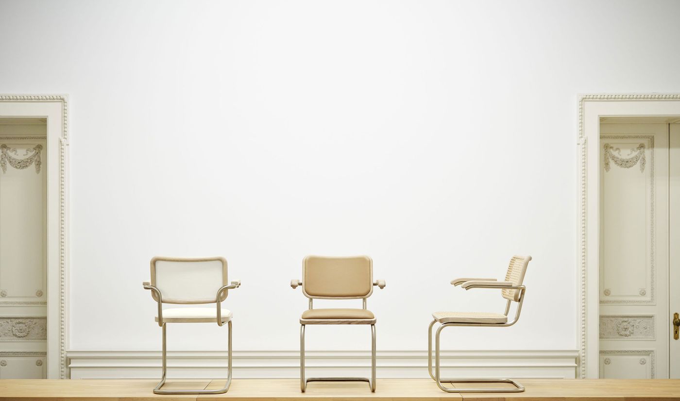 Three minimalist cantilever chairs in soft neutral tones, elegantly staged against an ornate, monochrome wall for a timeless contrast. Three minimalist cantilever chairs in soft neutral tones, elegantly staged against an ornate, monochrome wall for a timeless contrast.
