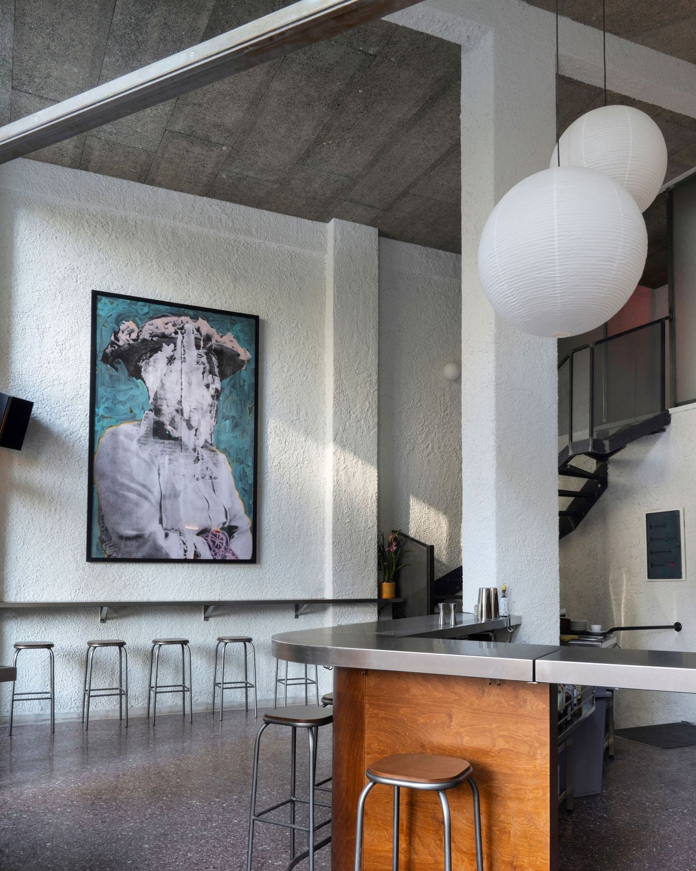 Inside Karate in Thessaloniki, a sculptural stainless-steel bar with timber panelling occupies a double-height interior characterised by textured white walls and exposed concrete ceilings. Oversized Japanese-style paper lanterns, minimalist metal stools and a large contemporary artwork introduce a distinctive interplay of industrial austerity and Japanese-inspired design.