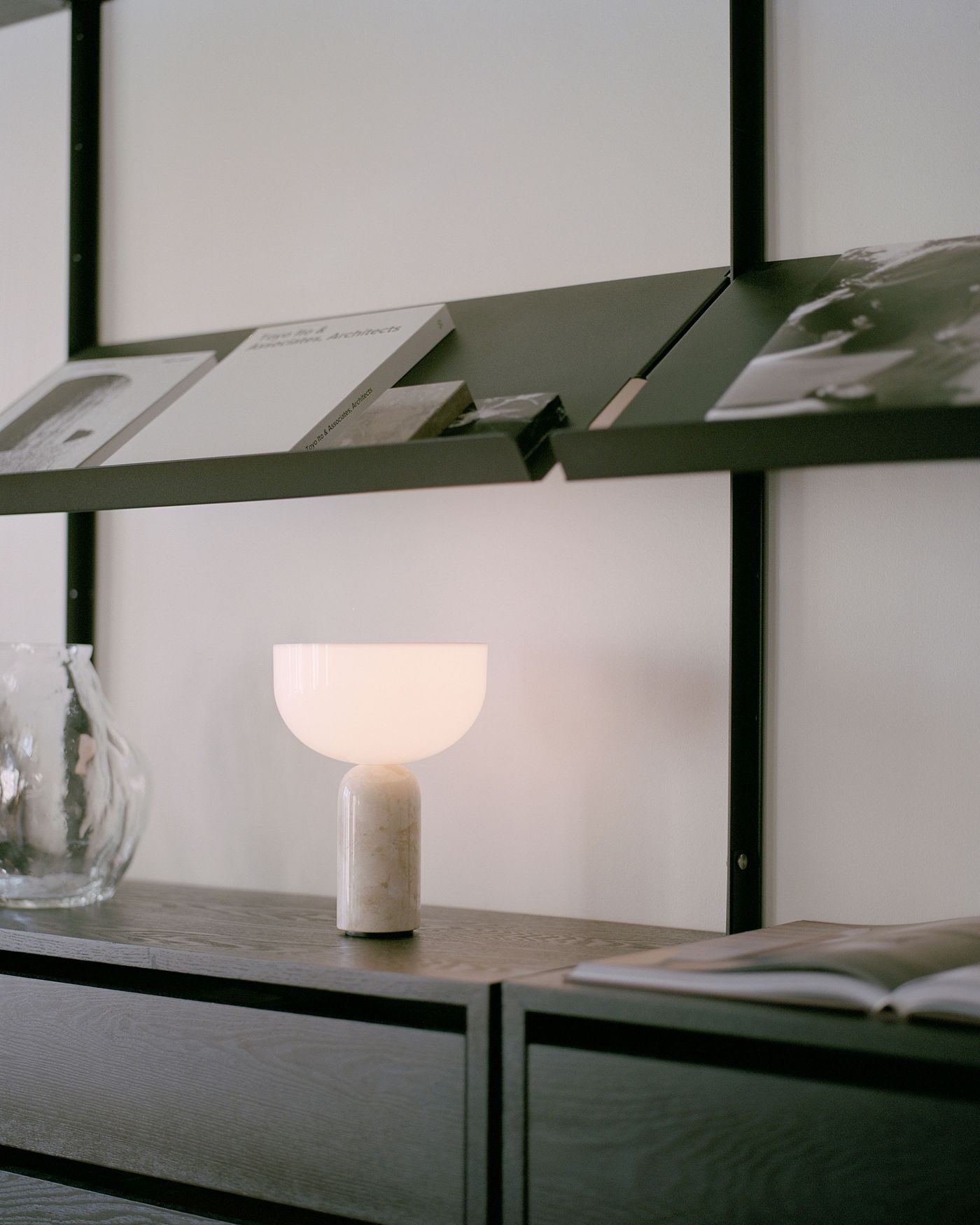 A softly glowing table lamp with a white, bowl-shaped shade and a cylindrical marble base sits on a dark wooden sideboard, next to a clear glass vase, with angled black shelves displaying books and magazines in the background. A softly glowing table lamp with a white, bowl-shaped shade and a cylindrical marble base sits on a dark wooden sideboard, next to a clear glass vase, with angled black shelves displaying books and magazines in the background.