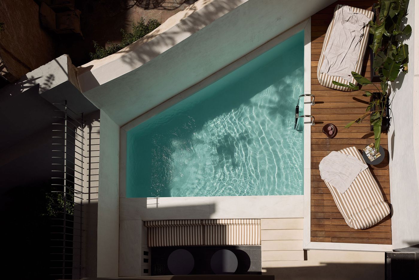Overhead view of a geometric outdoor swimming pool at Kuka in Athens, its turquoise water reflecting rippling sunlight. A narrow timber deck accommodates striped beige-and-white loungers, potted tropical greenery and small side tables. Crisp architectural lines, pale stone surfaces and deep shadows reinforce the secluded, contemporary Mediterranean atmosphere.